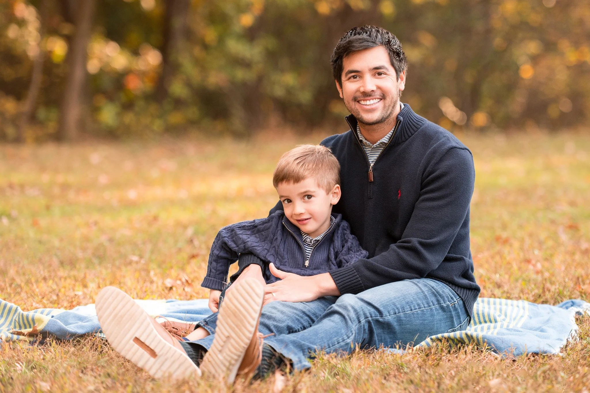 A father and son sitting on a blanket outdoors in a park during autumn. The father has dark hair and a beard, wearing a navy sweater, and is smiling. The young boy has light hair, wearing a dark blue jacket, and is leaning on his father, looking at t