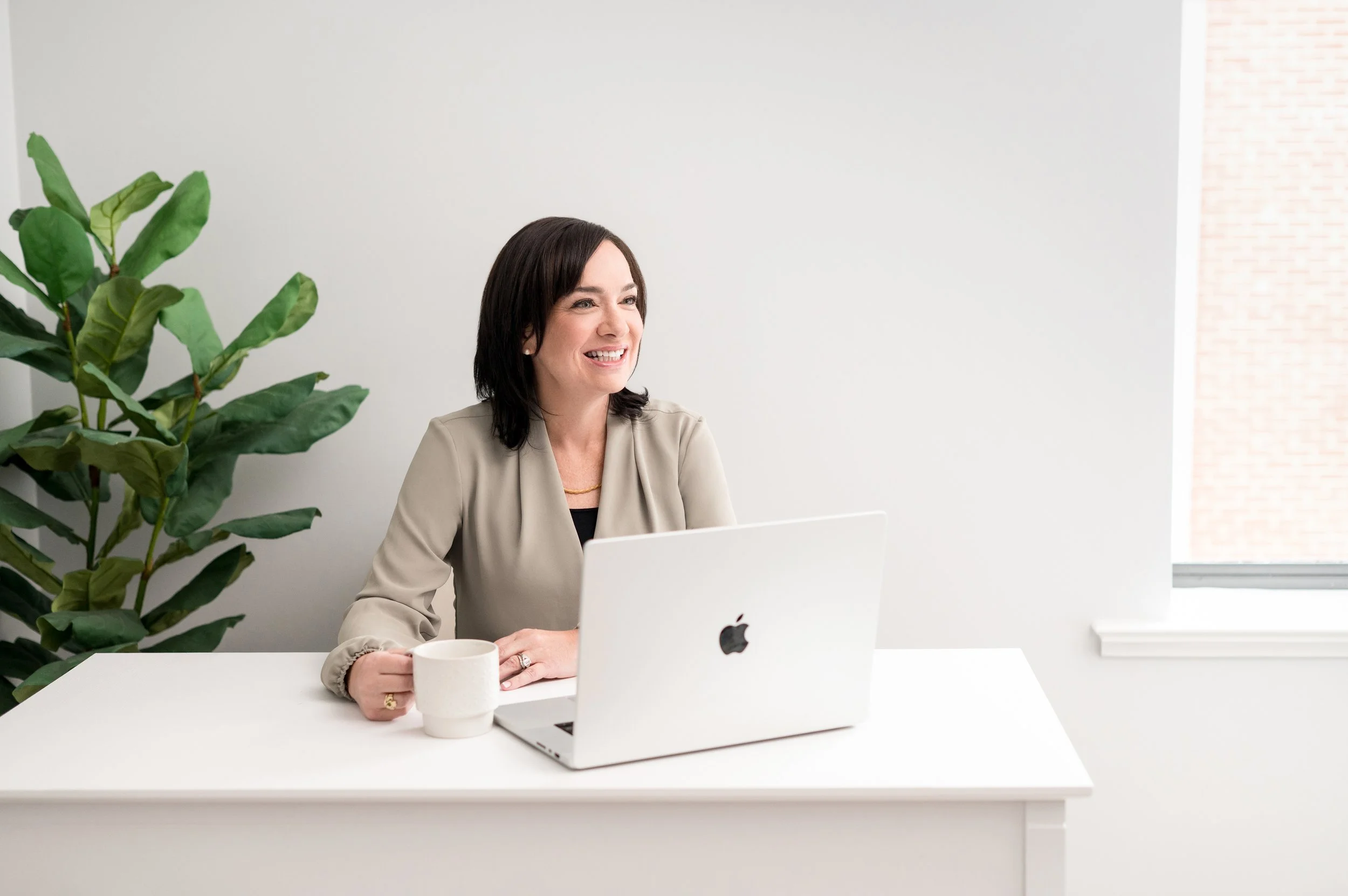 A woman with dark hair smiling while sitting at a white desk with a silver laptop and a white coffee mug in a bright room with a large green plant to her left and a window to her right.