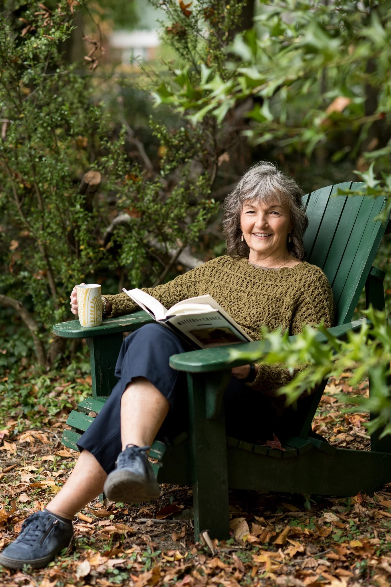 An elderly woman with gray hair, wearing a green knitted sweater and navy pants, sitting in a green Adirondack chair outdoors. She is smiling, holding a book in her lap, and a mug on the armrest. The setting is surrounded by lush green foliage and fa