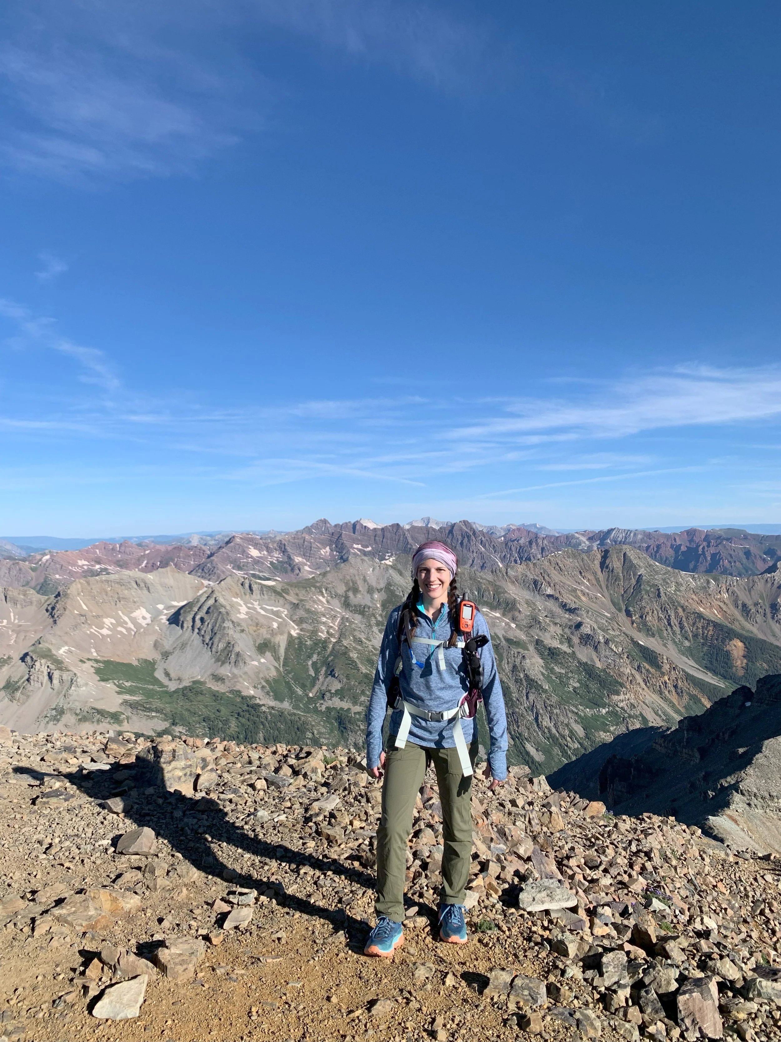 Katharine Moustakes on top of mountain or ridge with rocks afoot. She is dressed in cooler weather attire and has running vest on.