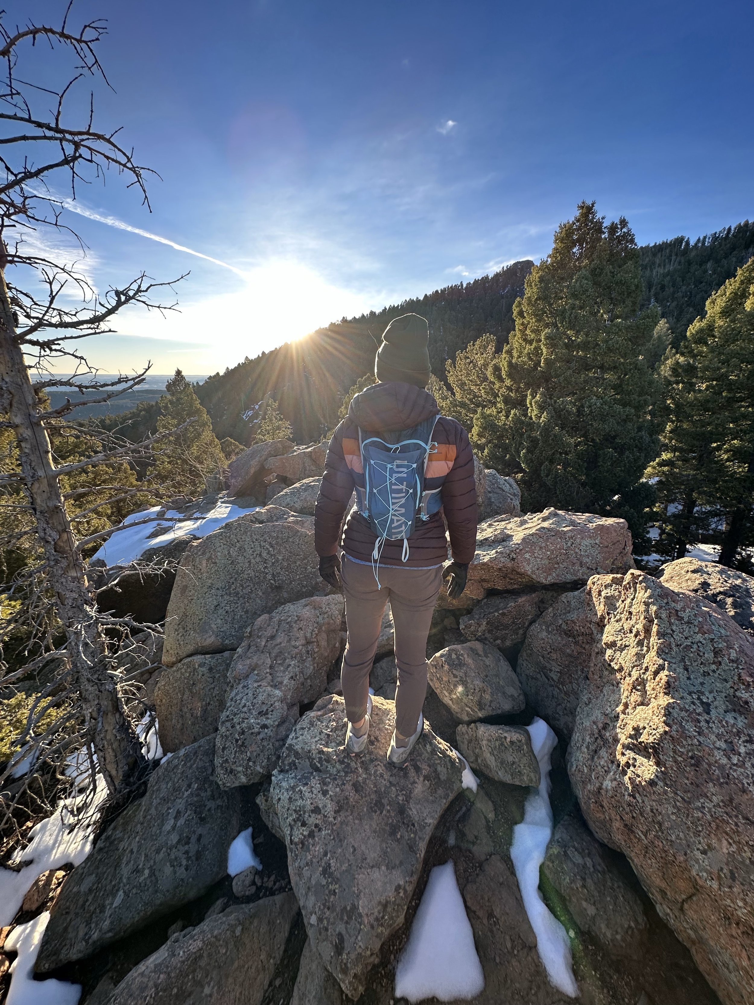 Picture of Katharine Moustakes from the back. Sun is peaking above the ridge in the background as Katharine is dressed in cooler weather attire, has a running best on, and is on boulders.