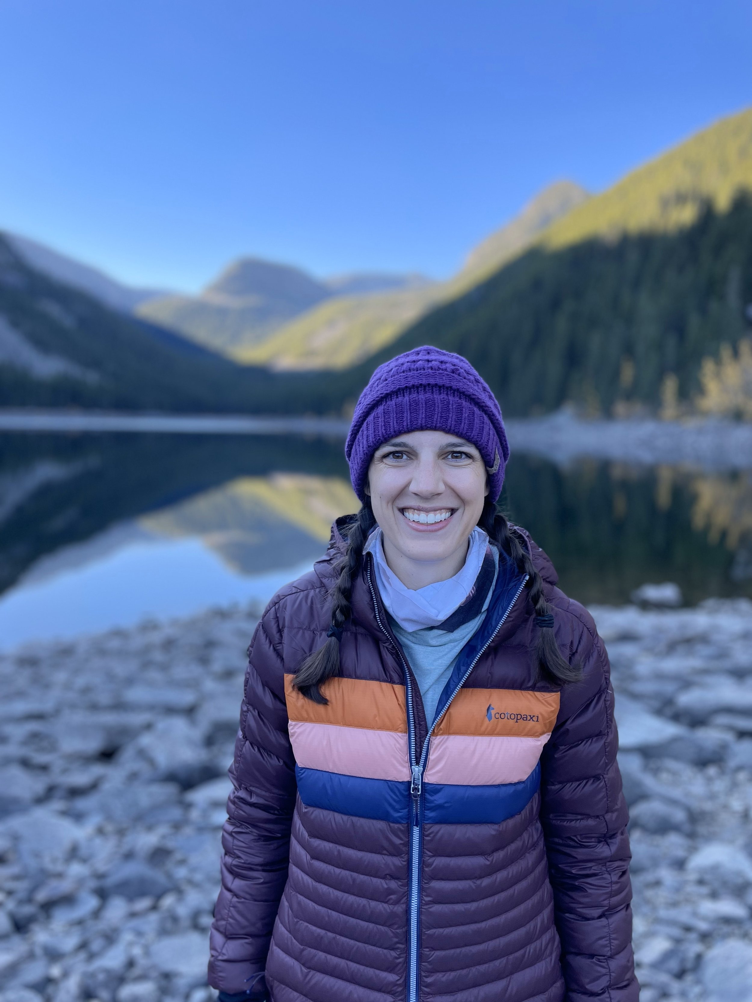 Picture of Katharine Moustakes wearing cooler weather attire and smiling in front of lake with Maroon Bells in the background.