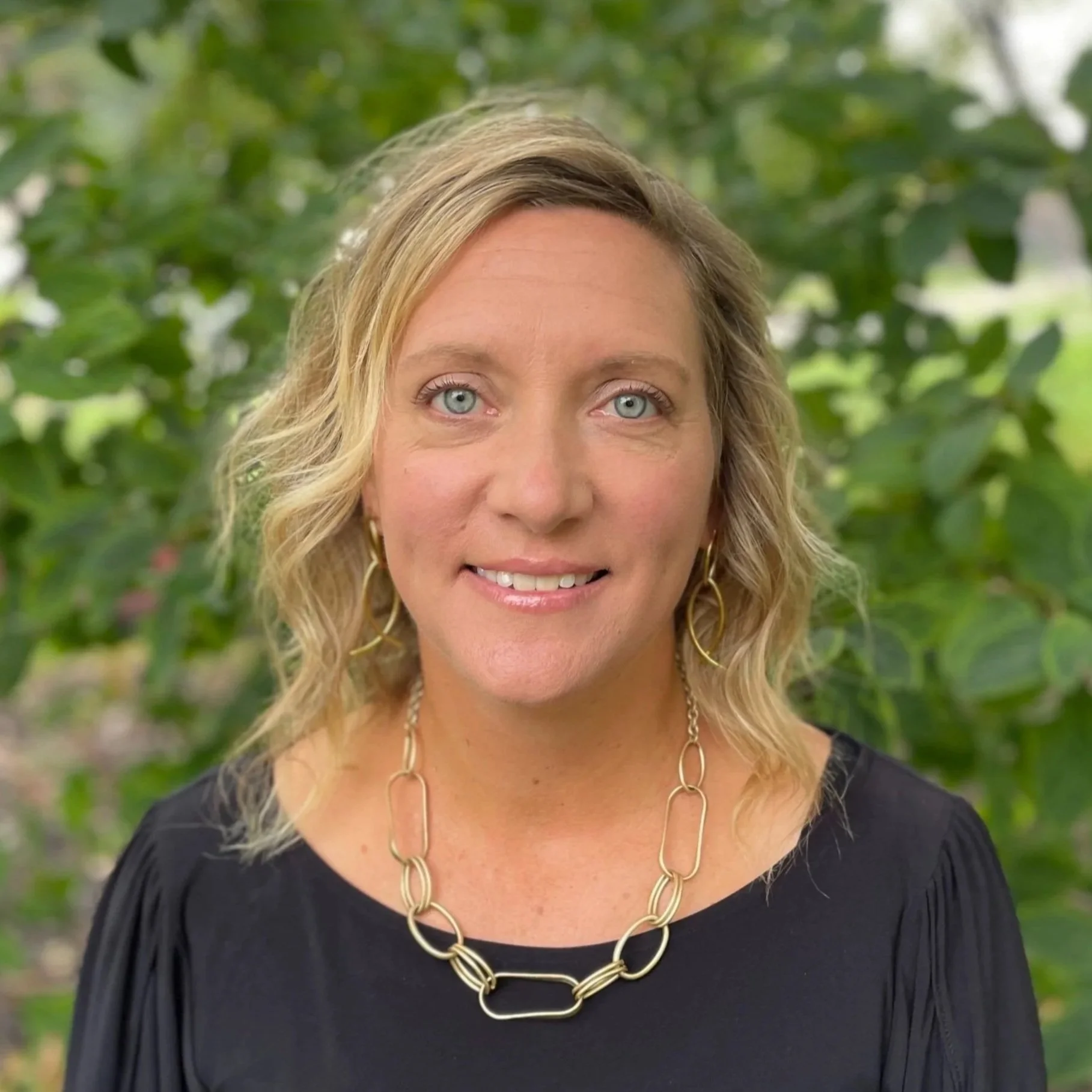 A woman with shoulder-length blonde hair, blue eyes, and wearing gold jewelry, standing outdoors with green foliage in the background.