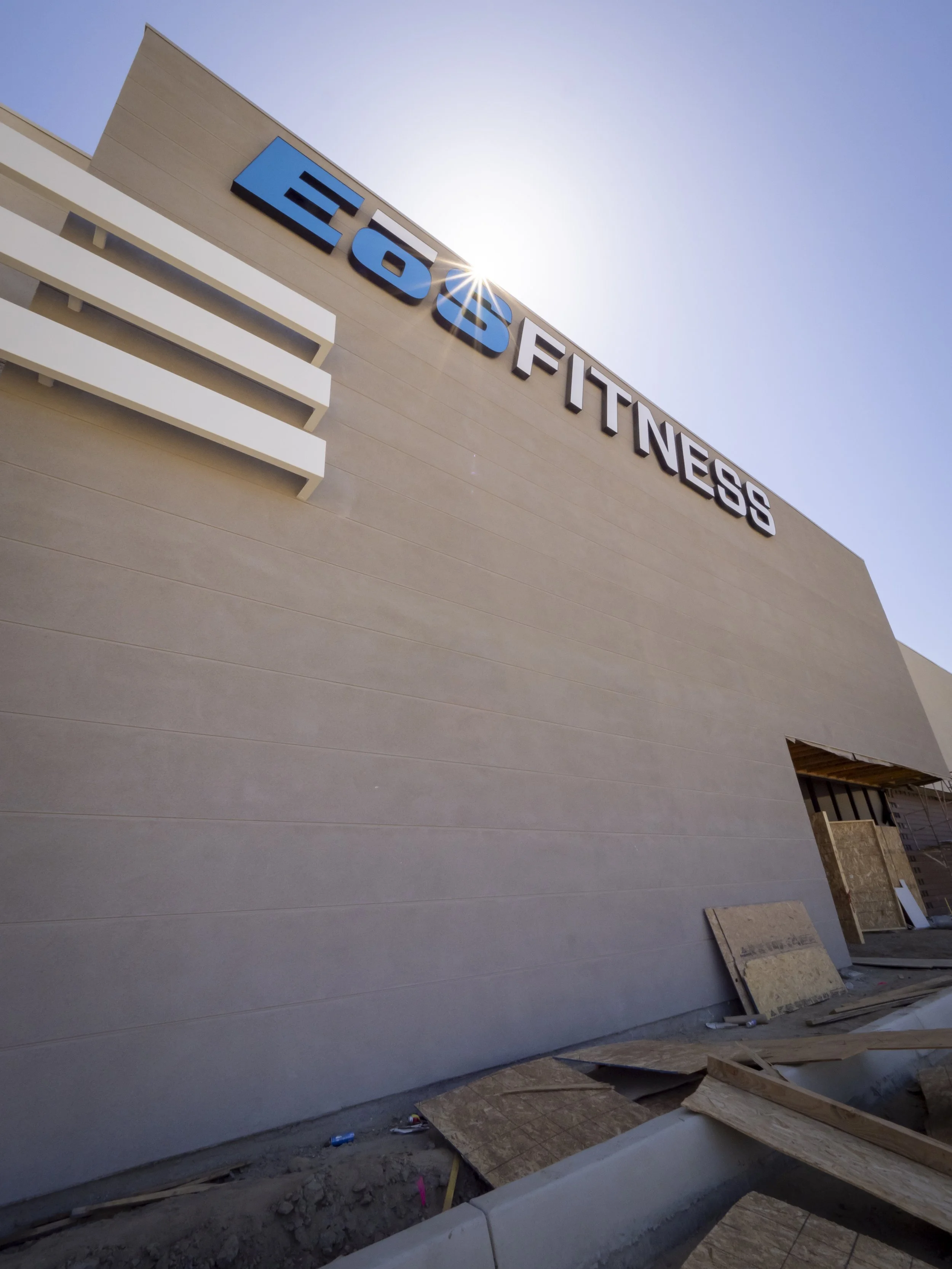 Exterior of a building under construction with a sign that reads 'EōB Fitness' on a beige wall, with construction materials and debris on the ground, and the sun shining above.