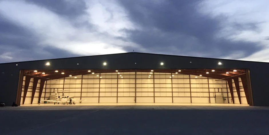 A large airplane hangar with a jet parked inside, illuminated at dusk with an open side showing the inside, under a cloudy sky.