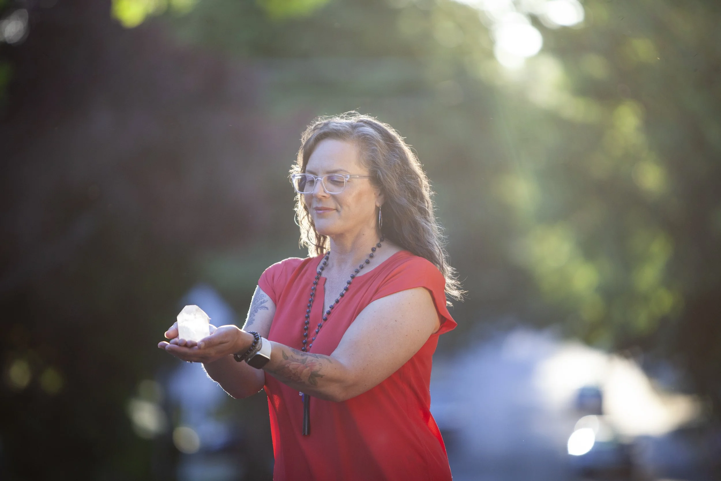 A woman with curly hair, glasses, and tattoos on her arms stands outdoors in sunlight, holding a white object in her hand.