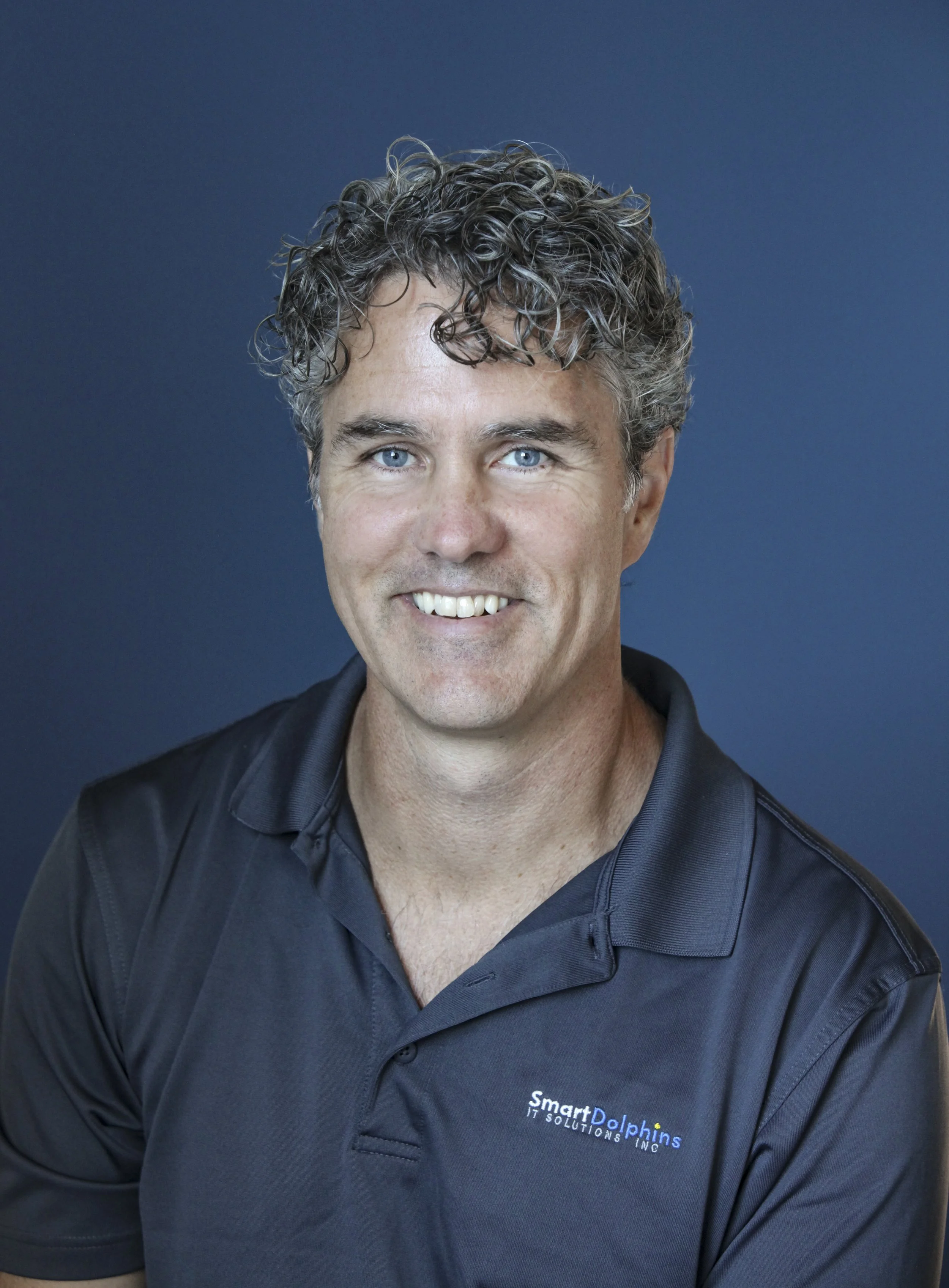 A smiling man with curly gray hair and blue eyes, wearing a black polo shirt with a logo for Smart Dolphins IT Solutions Inc, against a dark blue background.