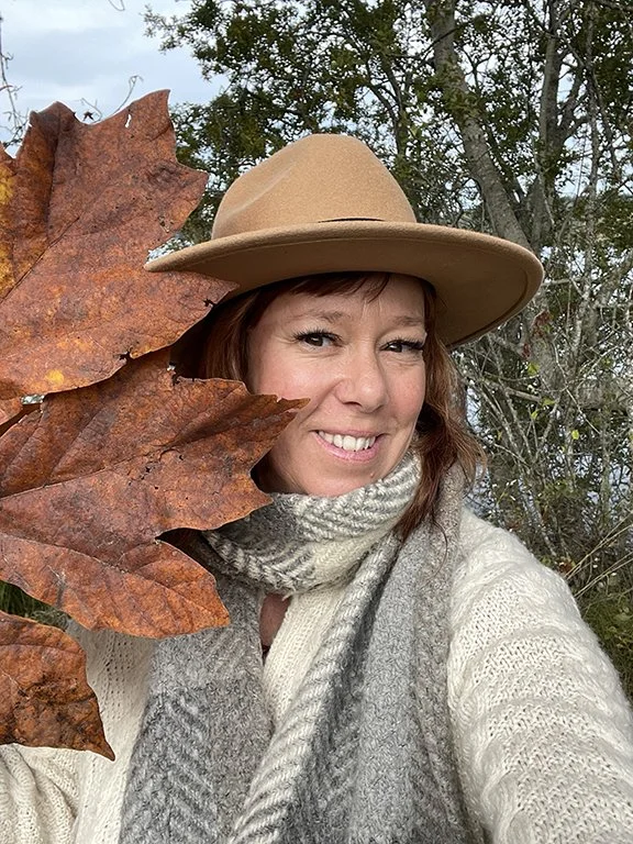 Woman wearing a beige hat and scarf, holding orange autumn leaves, smiling outdoors with trees in the background.