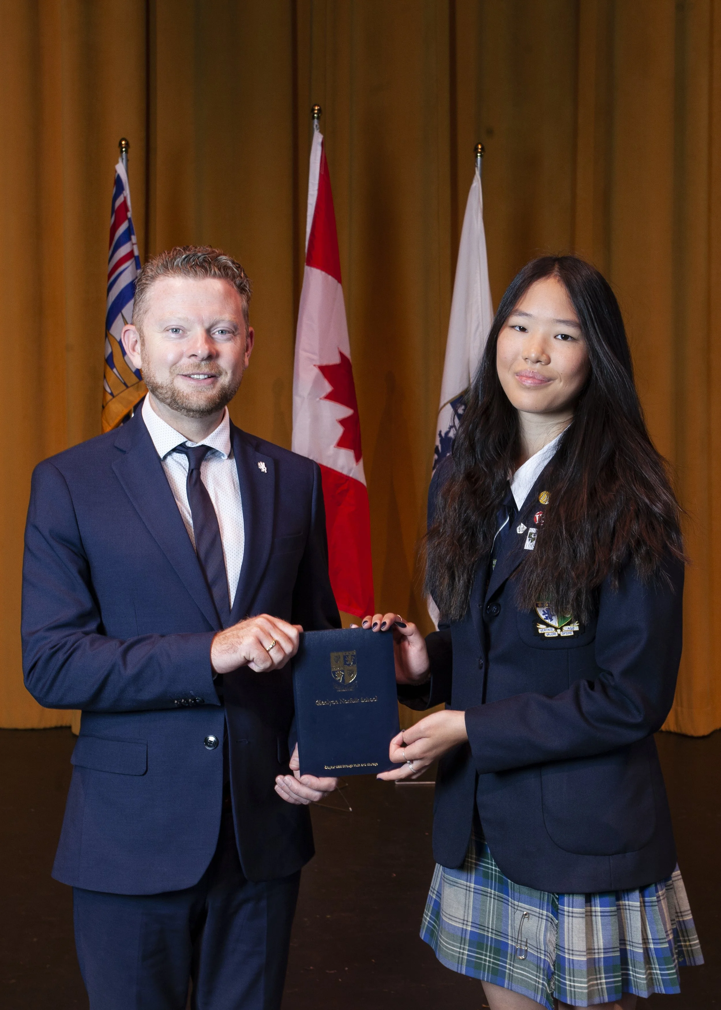 A young woman in a plaid skirt and blazer receiving a certificate from a man in a suit, in front of flags of British Columbia, Canada, and other entities, on a stage with yellow curtains.