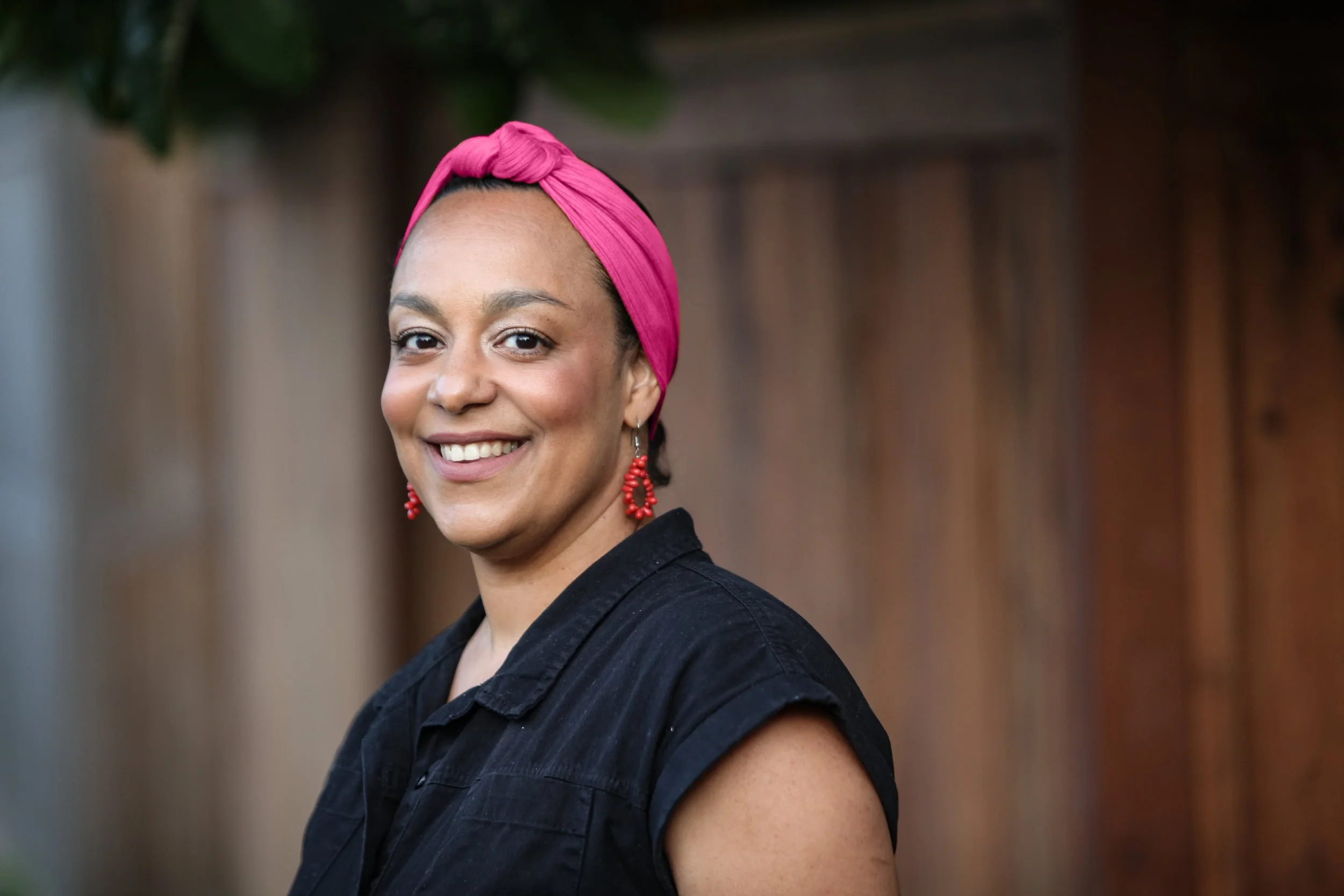 Woman smiling outdoors, wearing a black shirt, pink headscarf, and red earrings, with a wooden fence in the background.