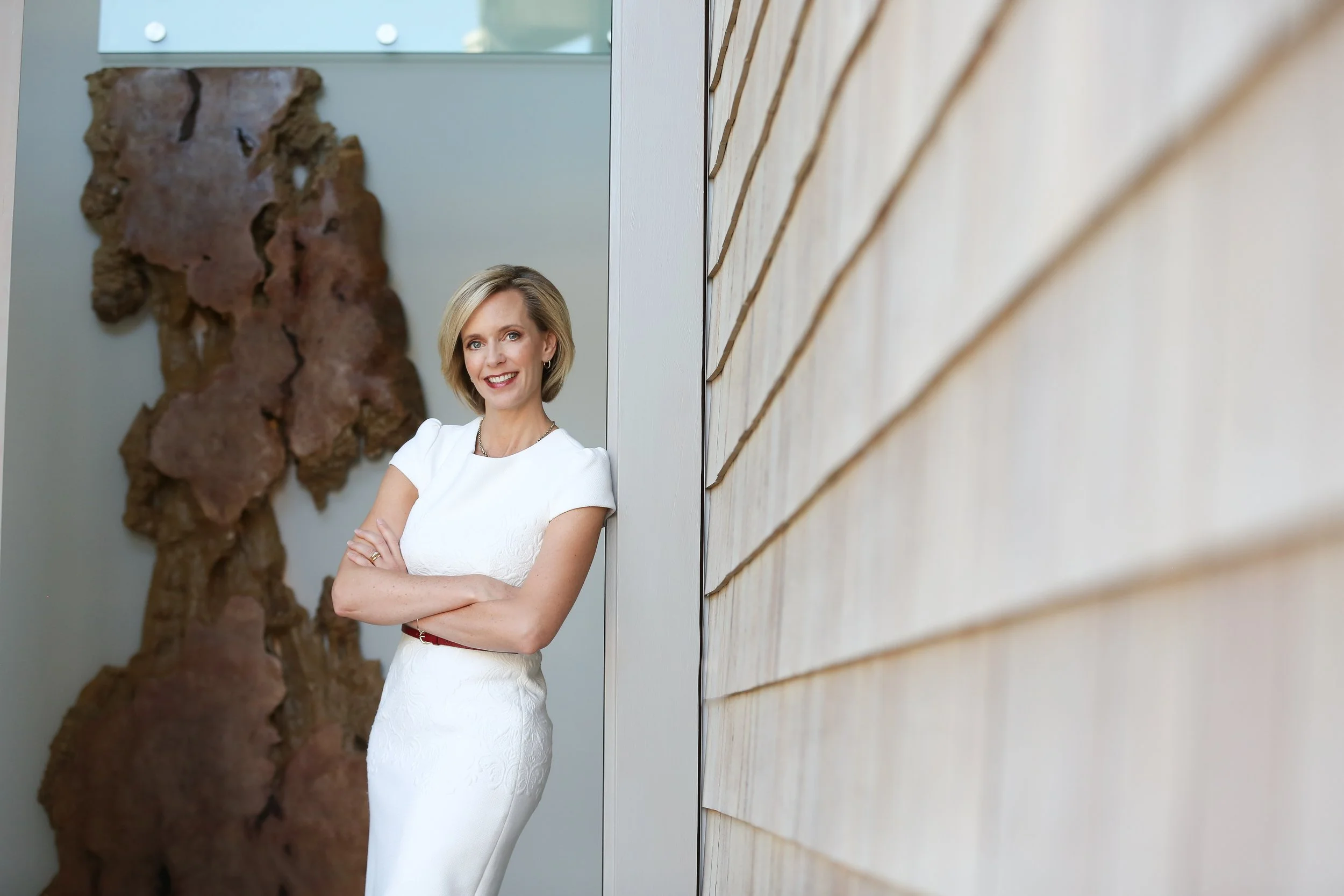 A woman with blonde hair wearing a white dress standing next to a wall with wooden siding, smiling with arms crossed.