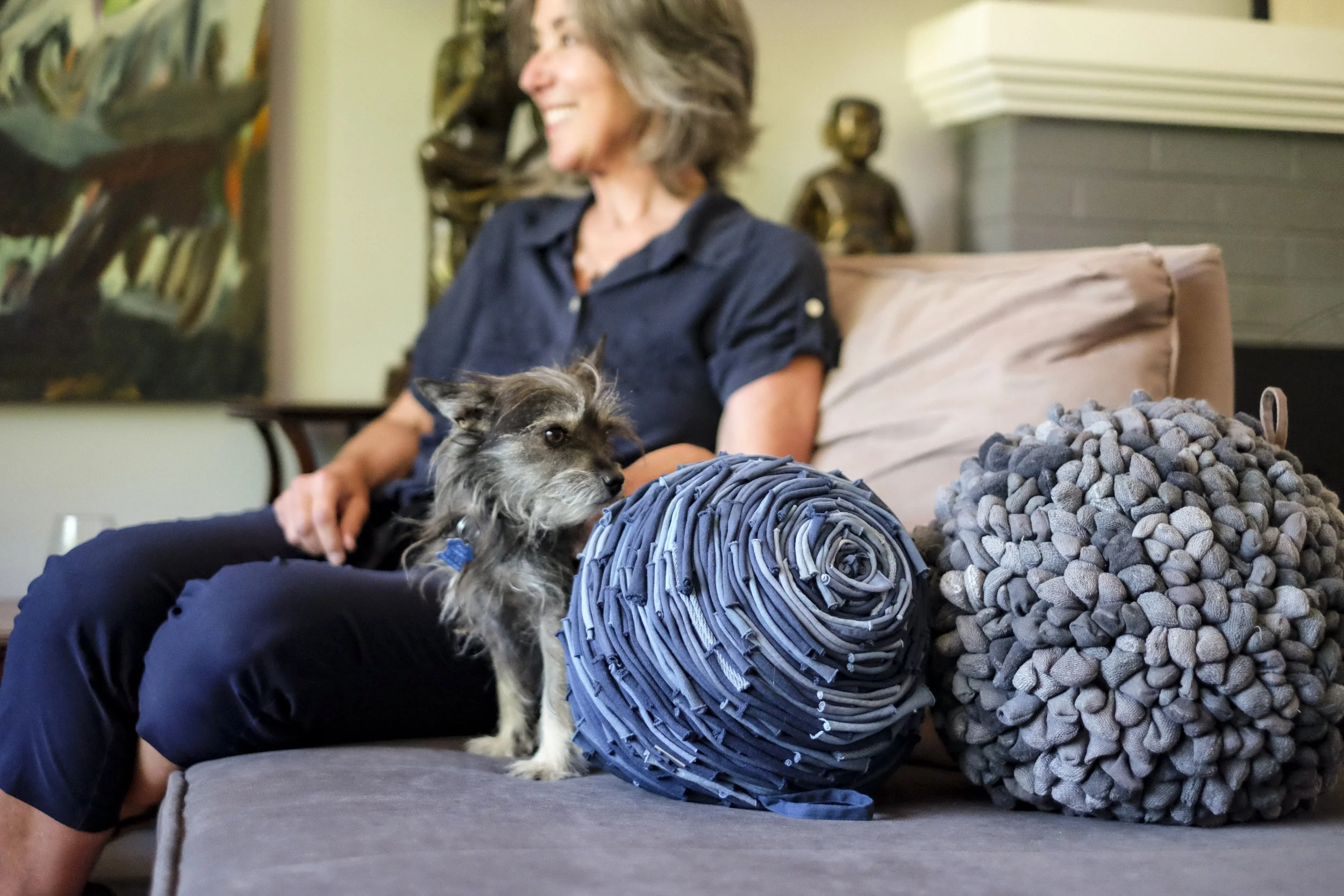 A woman sitting on a couch with a small gray and black dog next to her, looking off to the side. There are two large, textured gray pillows and a rolled-up fabric item on the couch. The background includes a piece of artwork and sculptures.