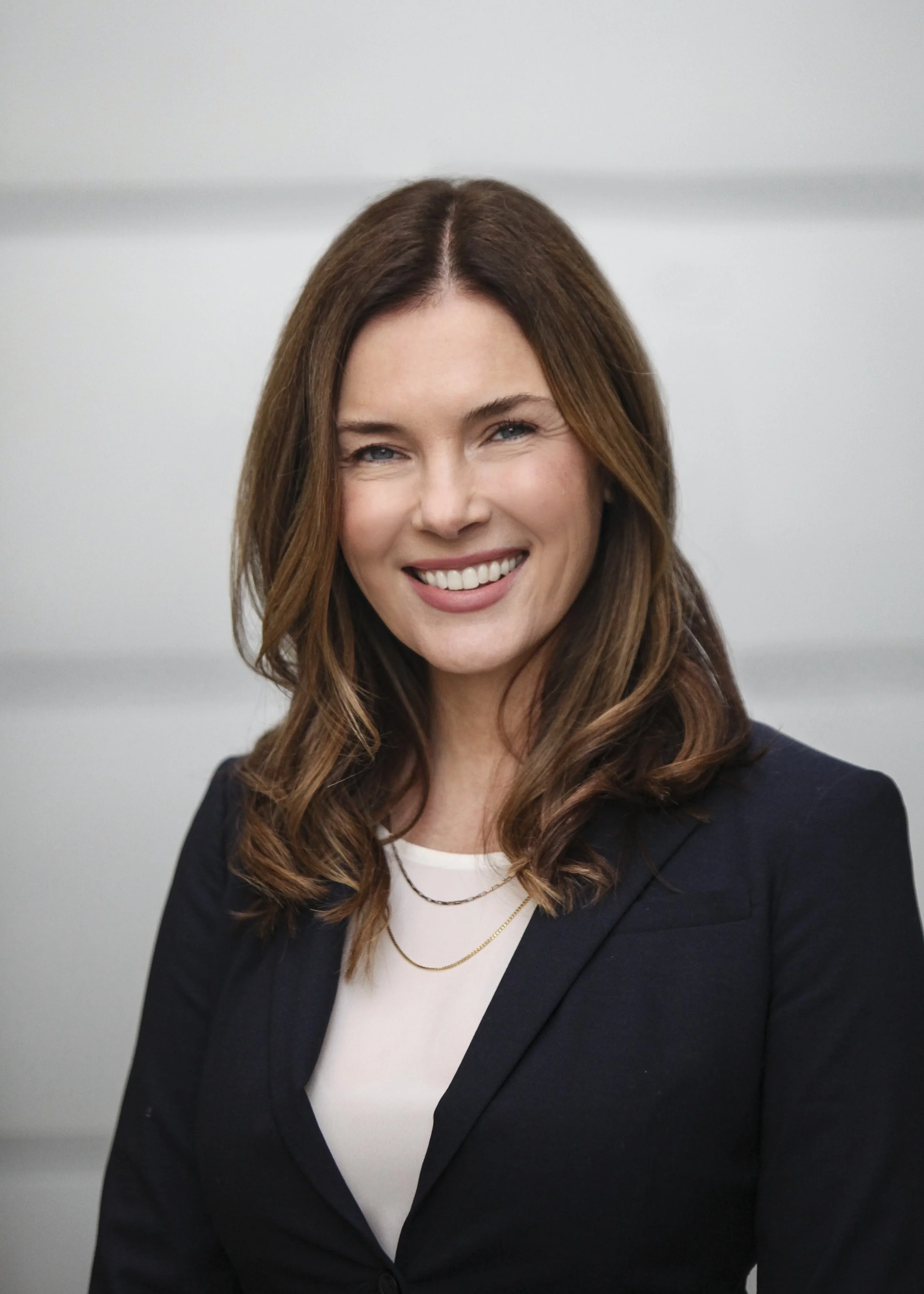 A woman with shoulder-length brown hair, wearing a black blazer over a white top, smiling in front of a blurred plain background.