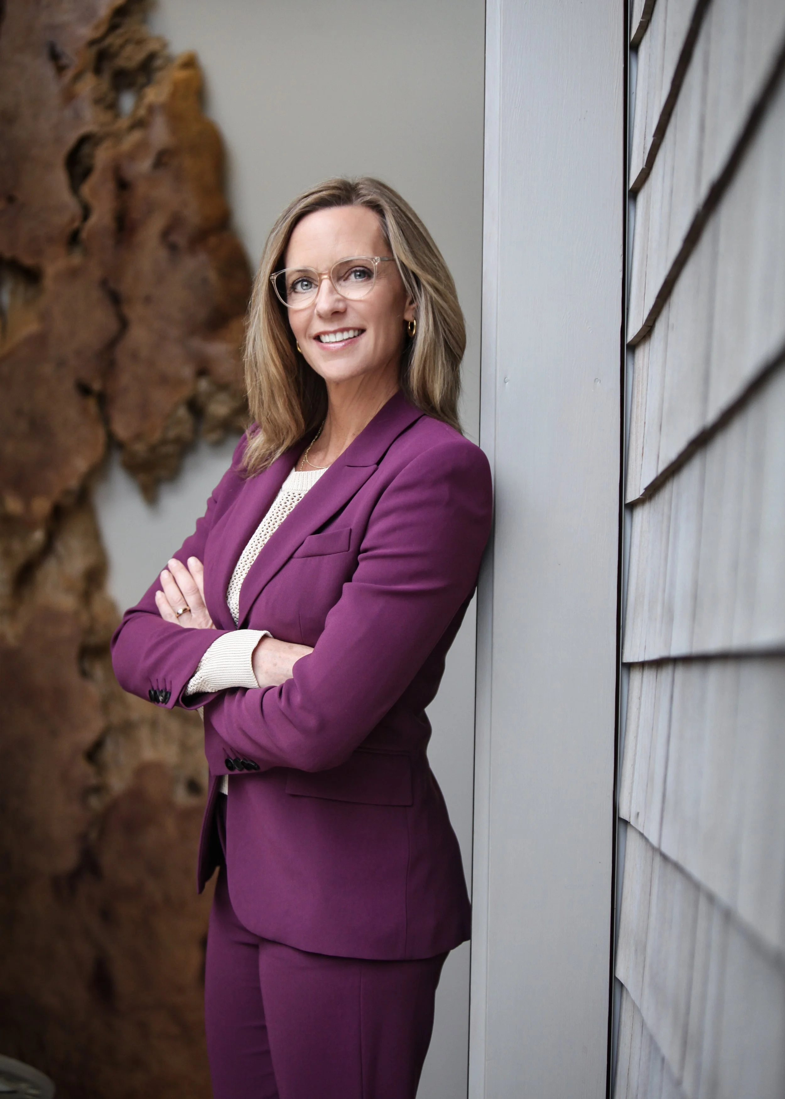 A woman with shoulder-length brown hair, wearing glasses, a purple suit, and a white blouse, standing with her arms crossed and smiling outdoors.