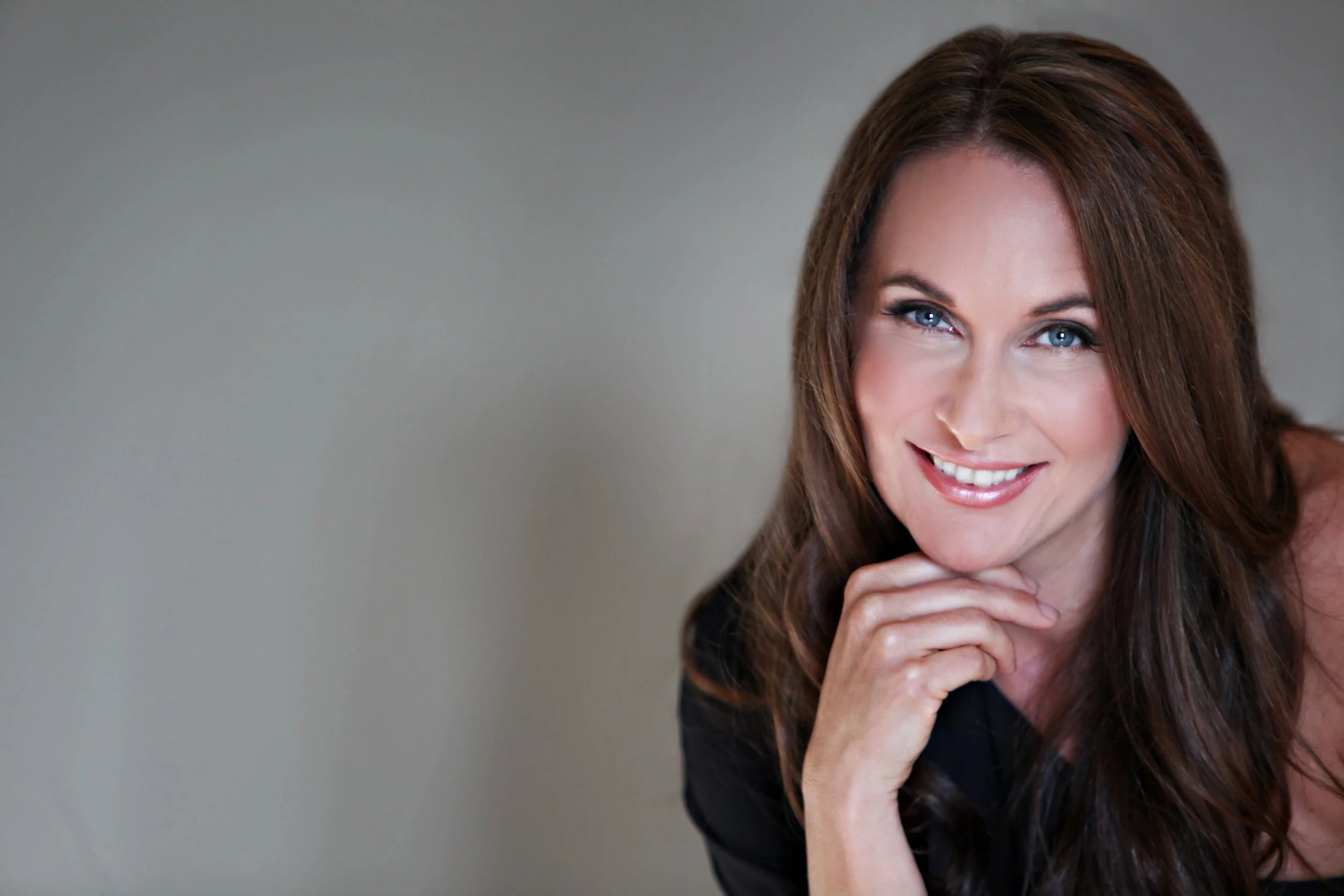 Close-up of a smiling woman with long brown hair and blue eyes, resting her chin on her hand against a plain background.