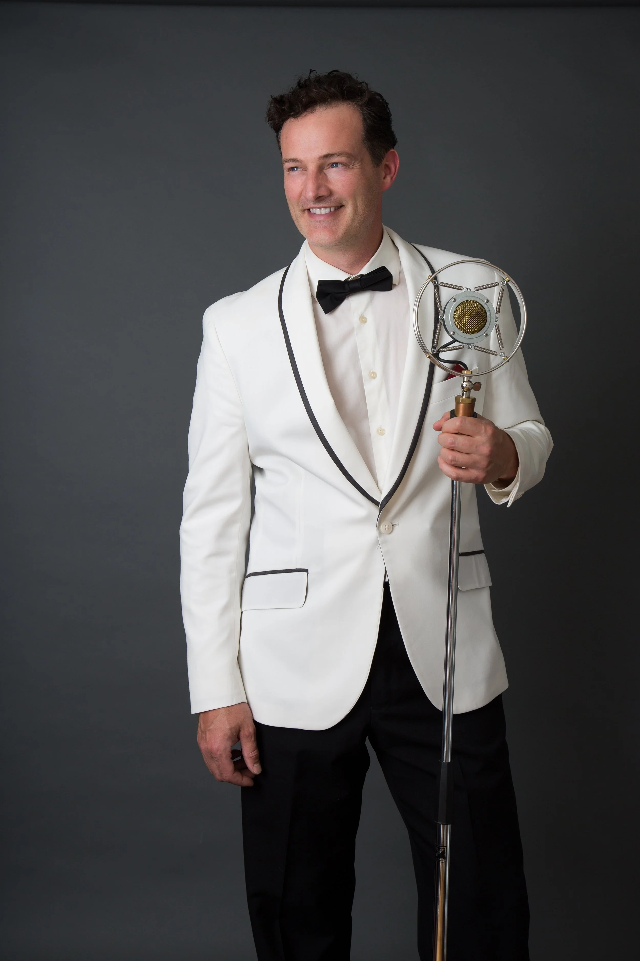 A man in a white tuxedo with black trim and a black bow tie holding a vintage microphone, standing against a dark background.