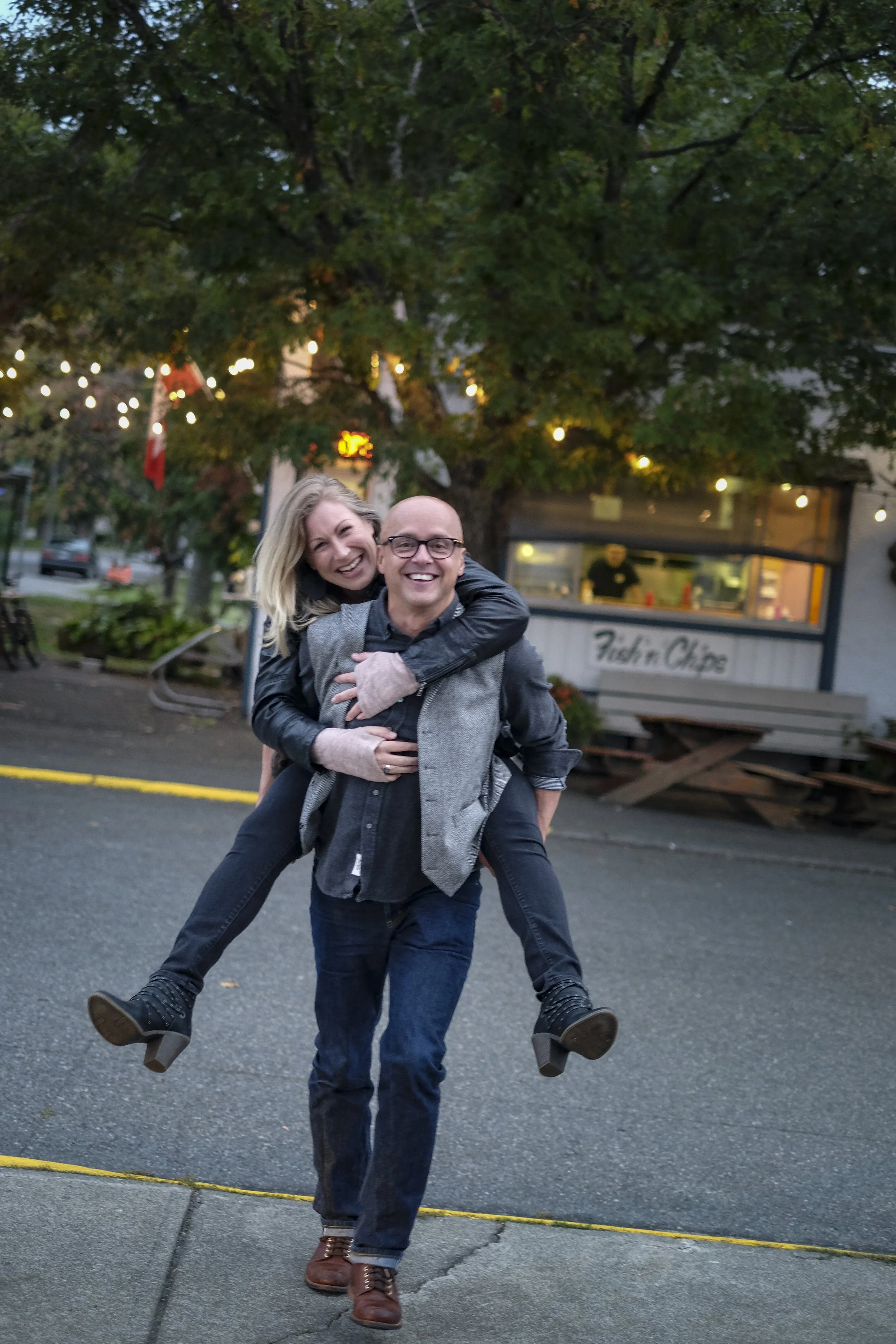 A man carrying a woman on his back outdoors in front of a tree with string lights and a food stand that says 'Fish'n Chips'. Both are smiling and appear happy.