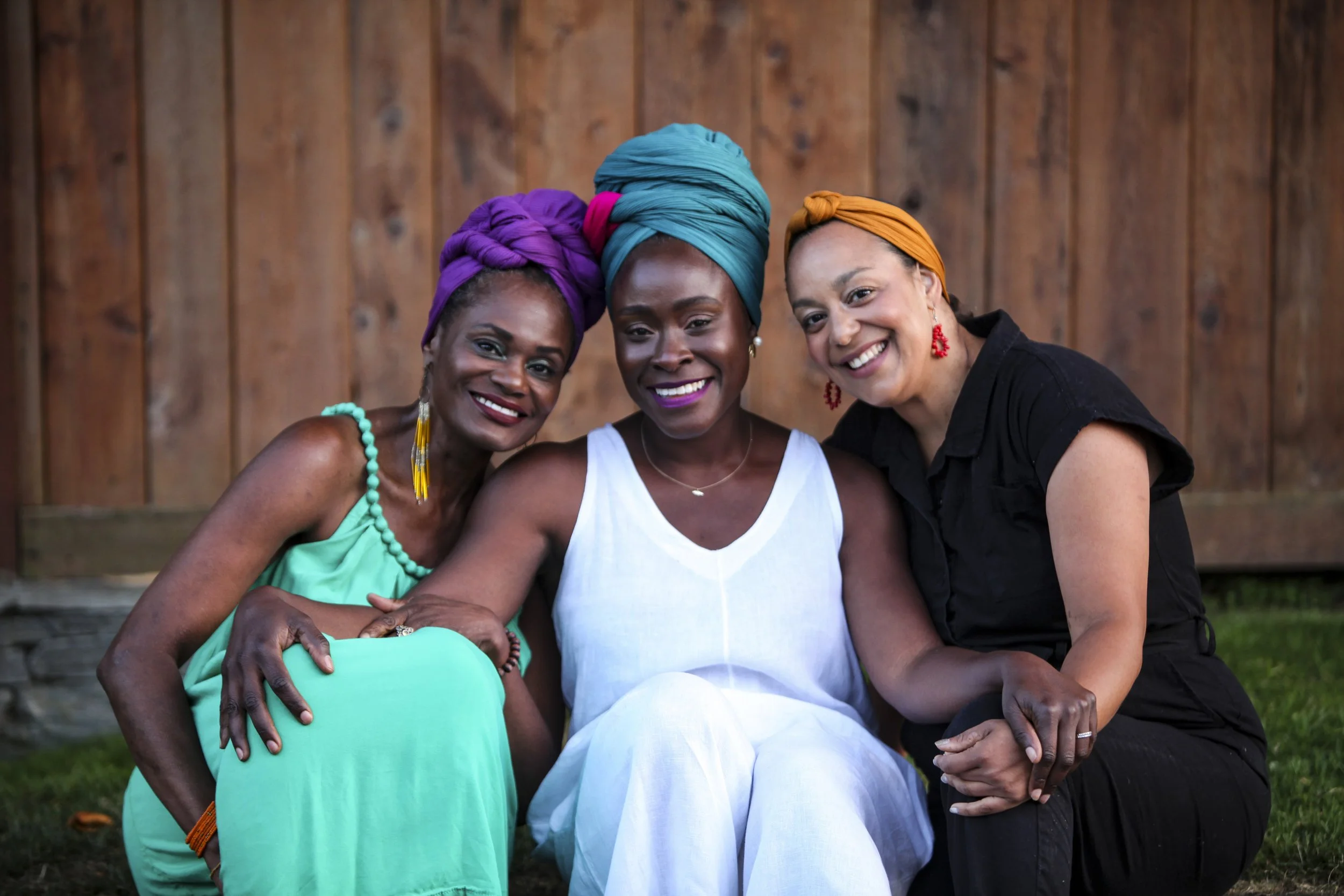 Three women sitting outdoors in front of a wooden fence, smiling and holding hands, wearing festive headwraps and colorful clothing.
