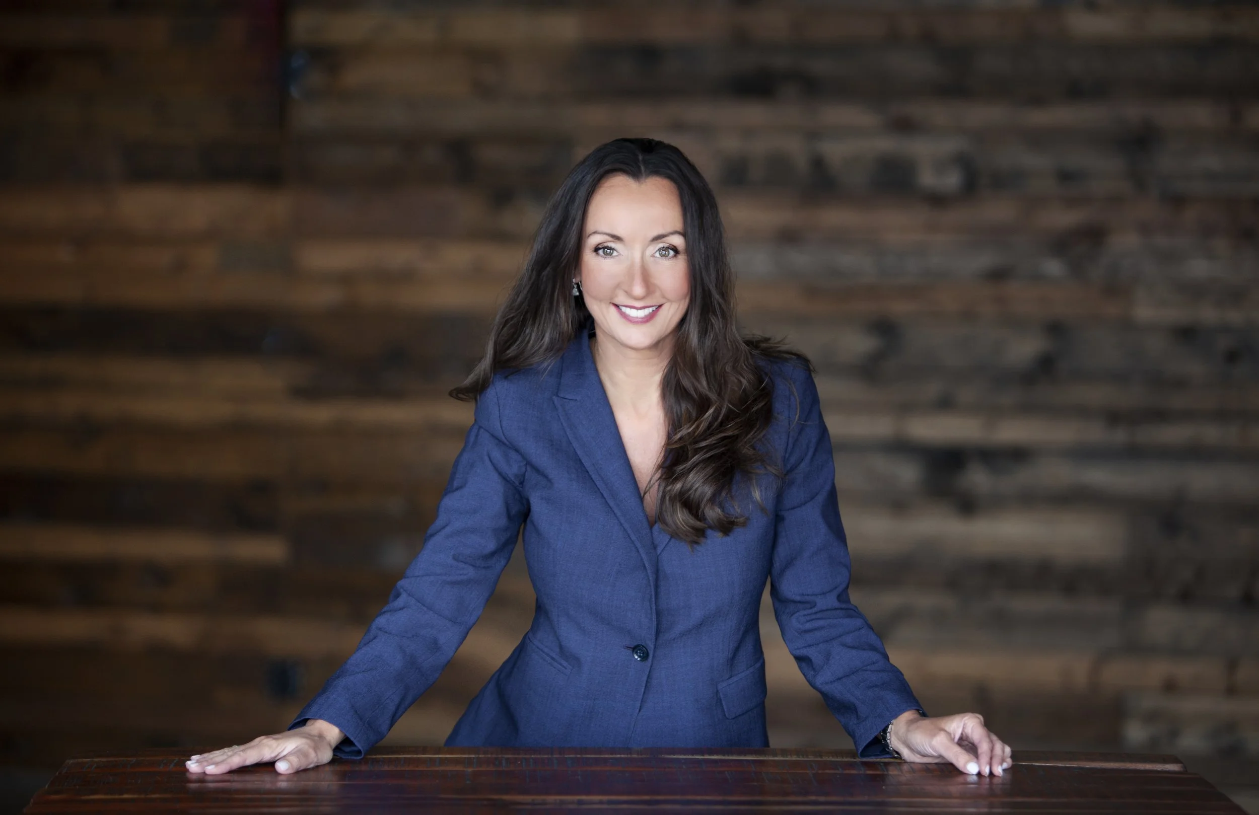 A woman with long brown hair, wearing a blue blazer, leaning on a wooden table and smiling at the camera against a wood-paneled background.
