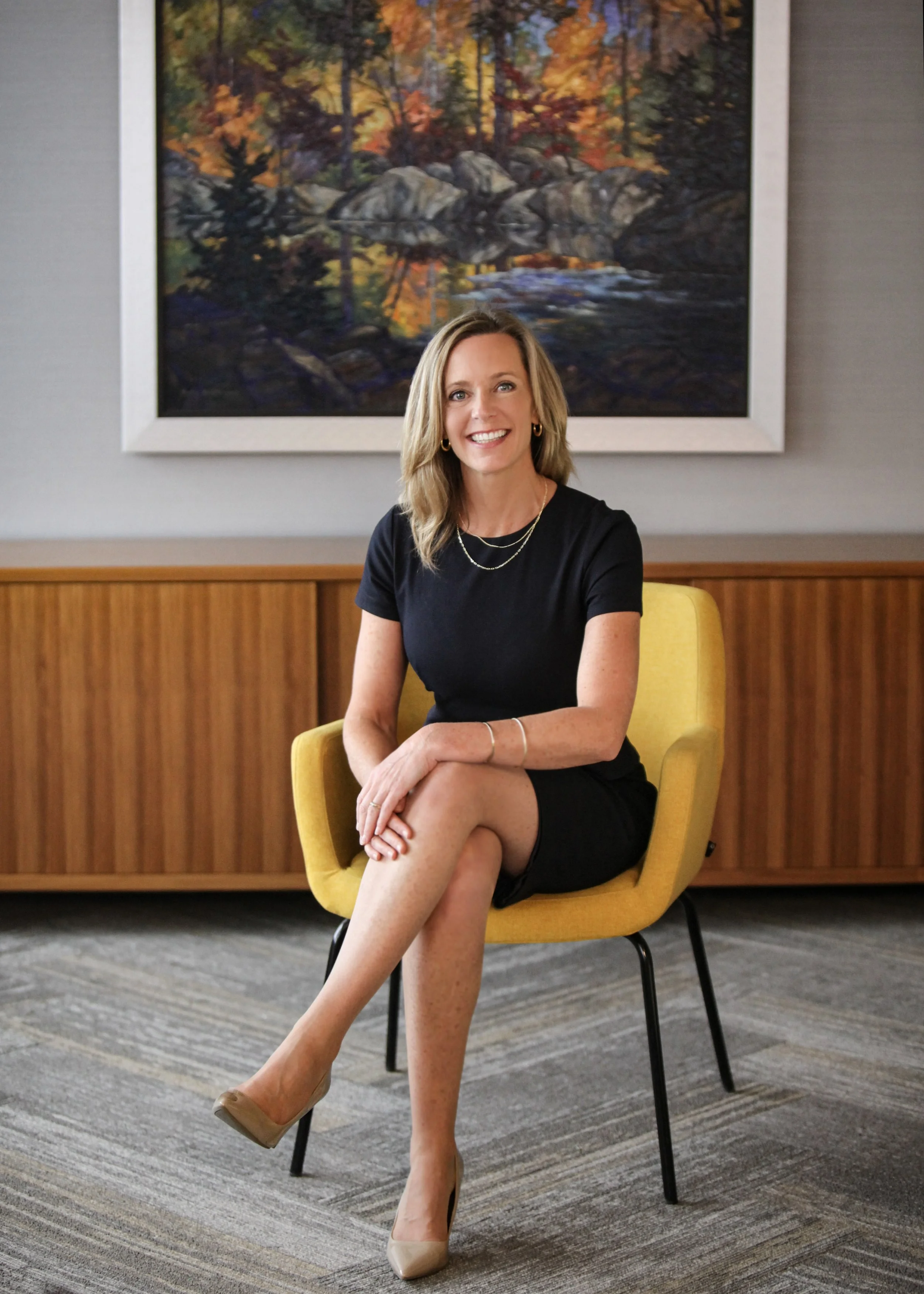 A woman sitting on a yellow chair in an office setting, smiling, with a large framed landscape painting of a forest scene behind her.