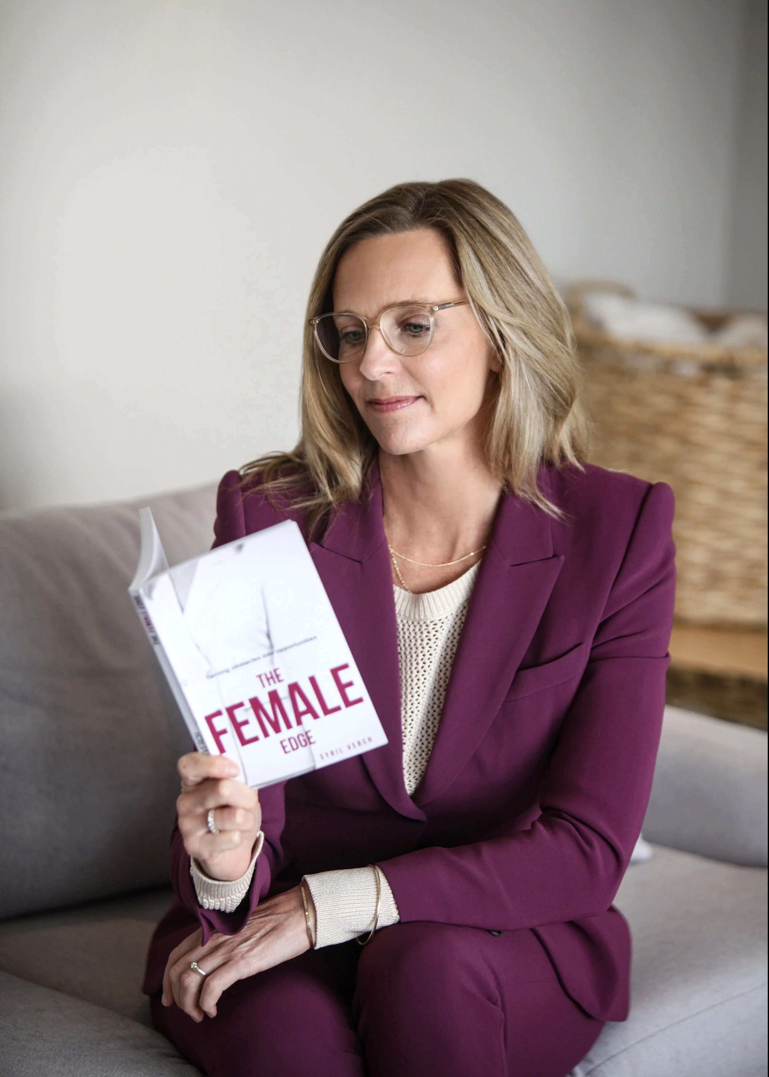 A woman with blonde hair, glasses, and wearing a purple blazer and pants, sitting on a grey couch, reading a book titled 'The Female Edge' by Sydnee Tucker.