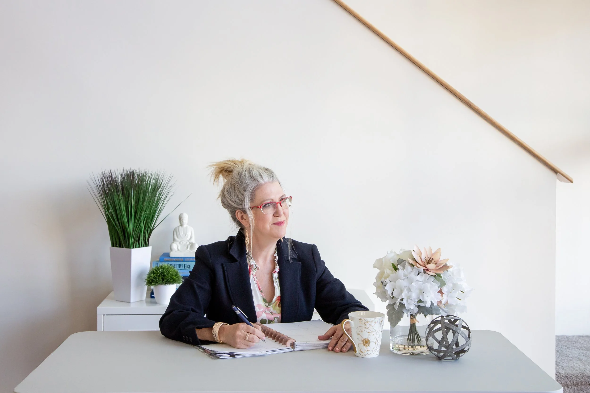 A woman with gray hair, glasses, and a black blazer sitting at a white desk with a notebook, a pen, a floral mug, and decorative objects including a white flowers bouquet and two spherical sculptures.