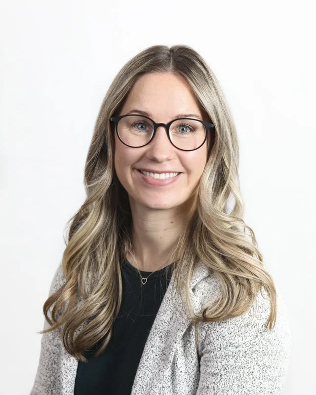 A woman with long wavy blonde hair, wearing glasses, a light grey blazer, and a black top, smiling at the camera against a plain white background.