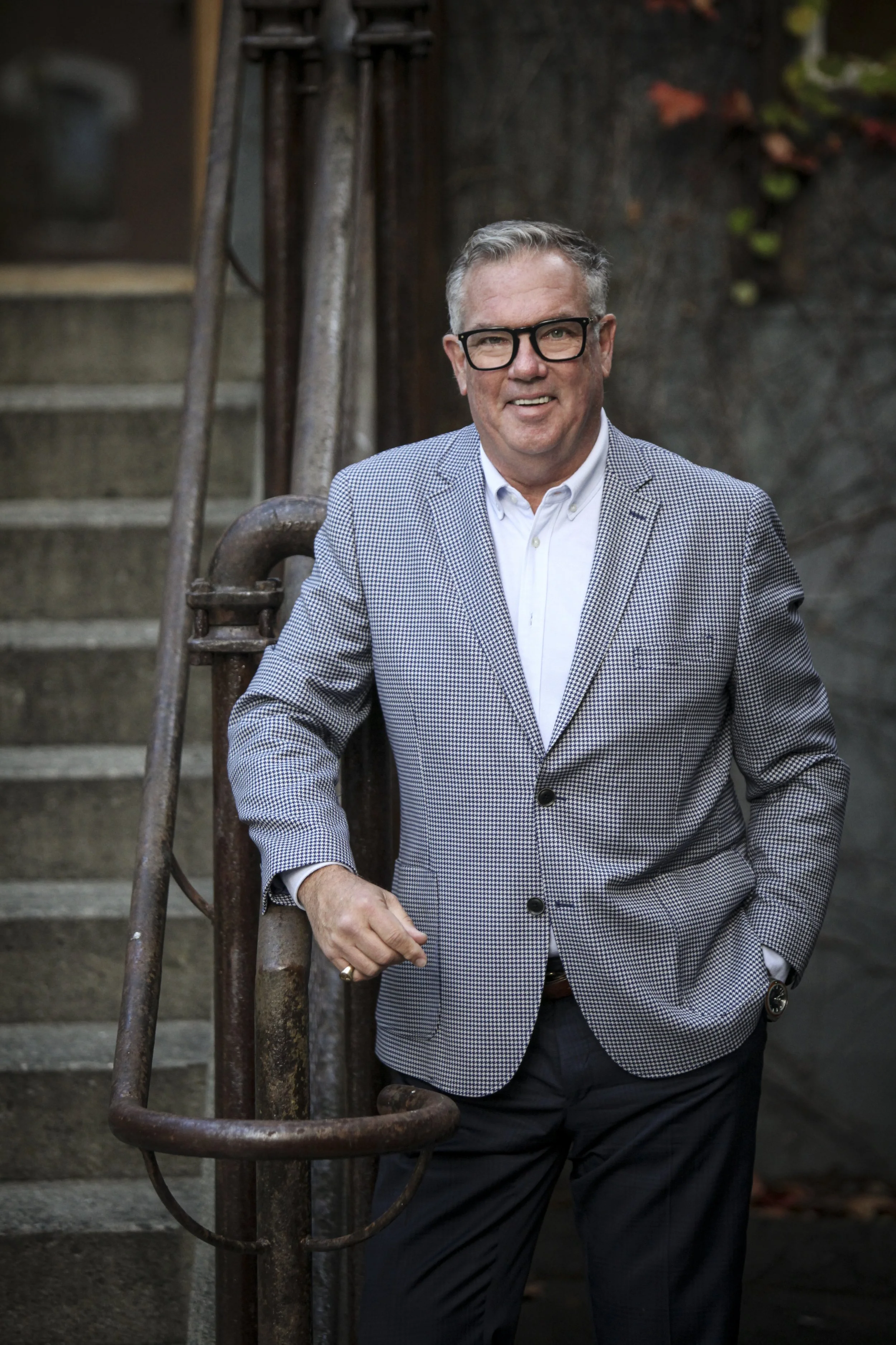 Man in a checkered blazer, white shirt, black glasses, standing outdoors near a metal railing and concrete stairs.