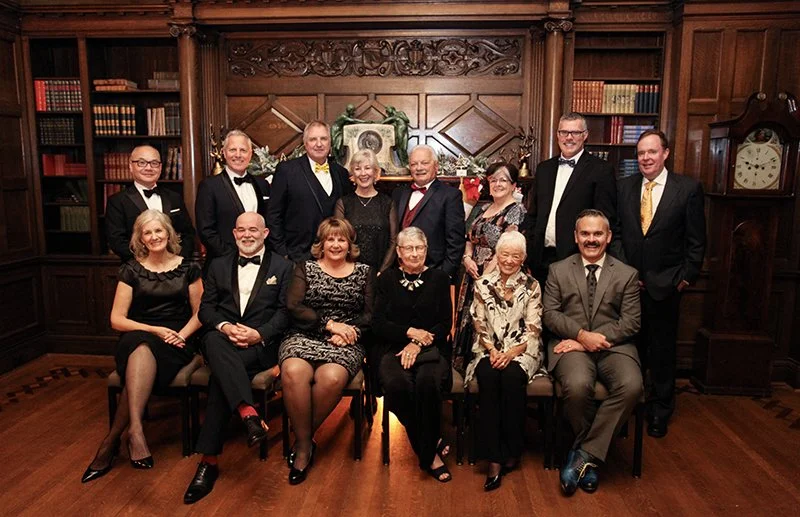 Group portrait of 14 people, mostly in formal attire, posing in a wood-paneled room with bookshelves, fireplace, and clock, for a family or special event.