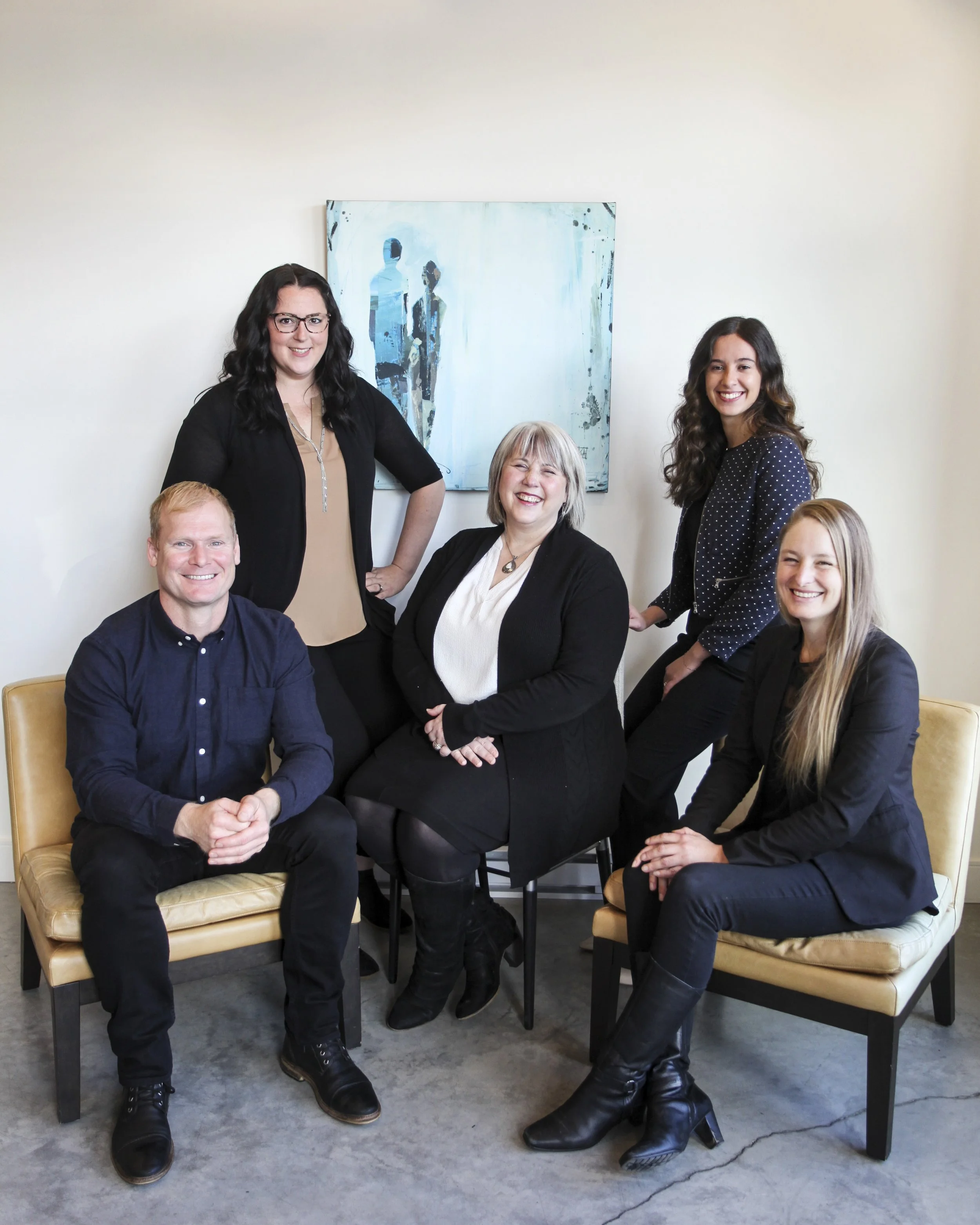 Group portrait of five smiling people in an office, with a painting of abstract figures on the wall behind them.