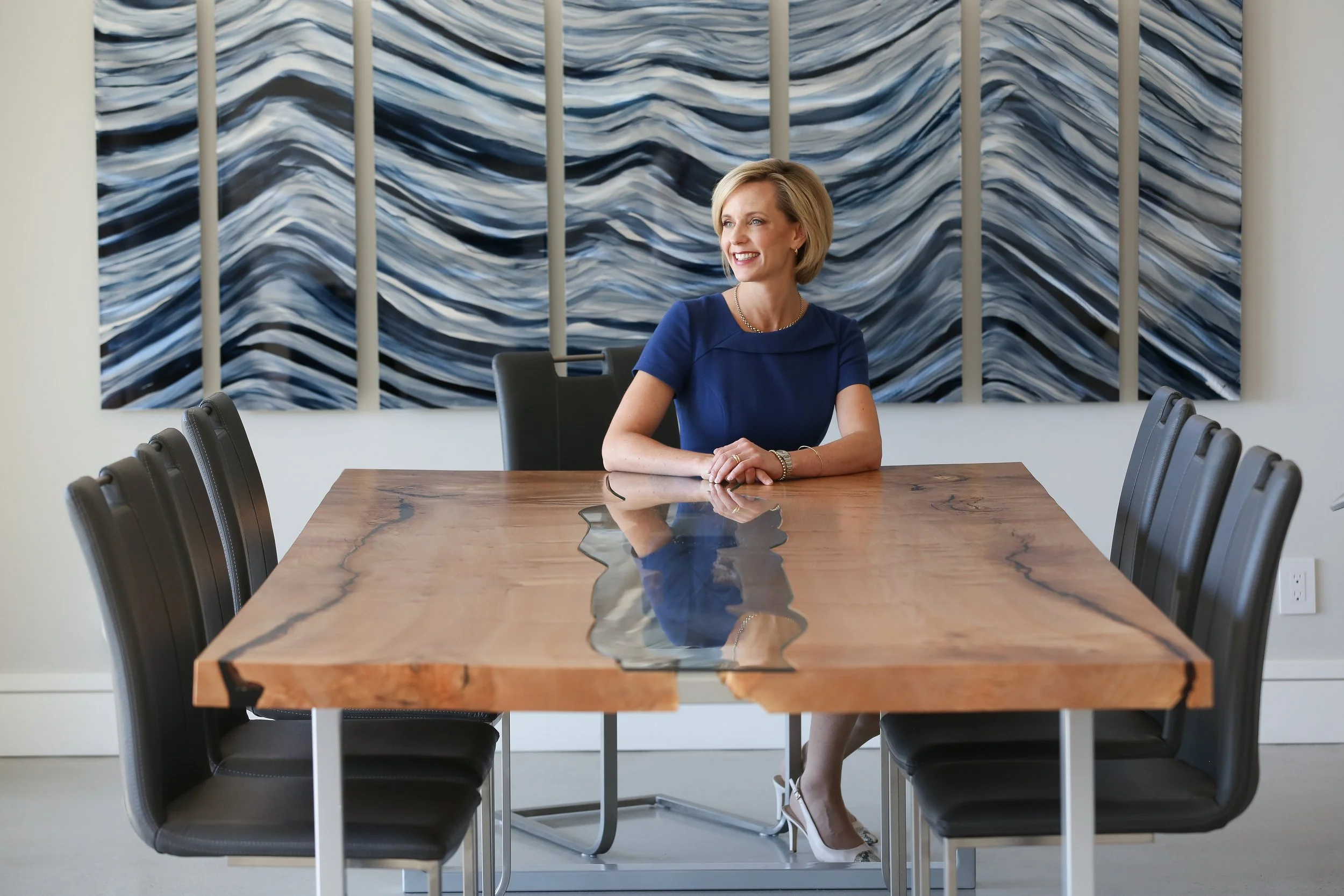 A woman in a blue dress sitting at a wooden conference table with black chairs, with a wavy abstract painting behind her.