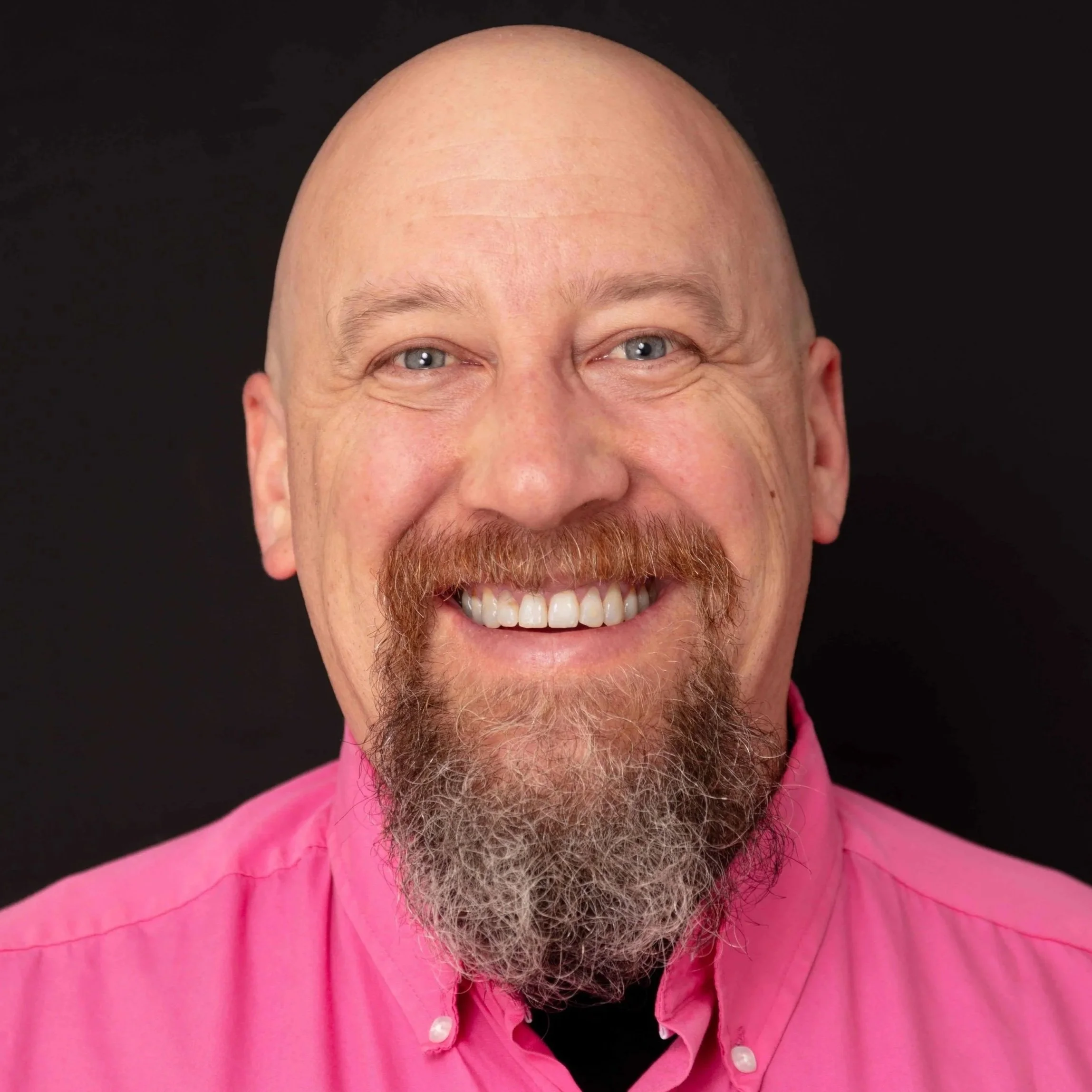 A smiling man with a bald head and a beard, wearing a bright pink collared shirt, against a black background.