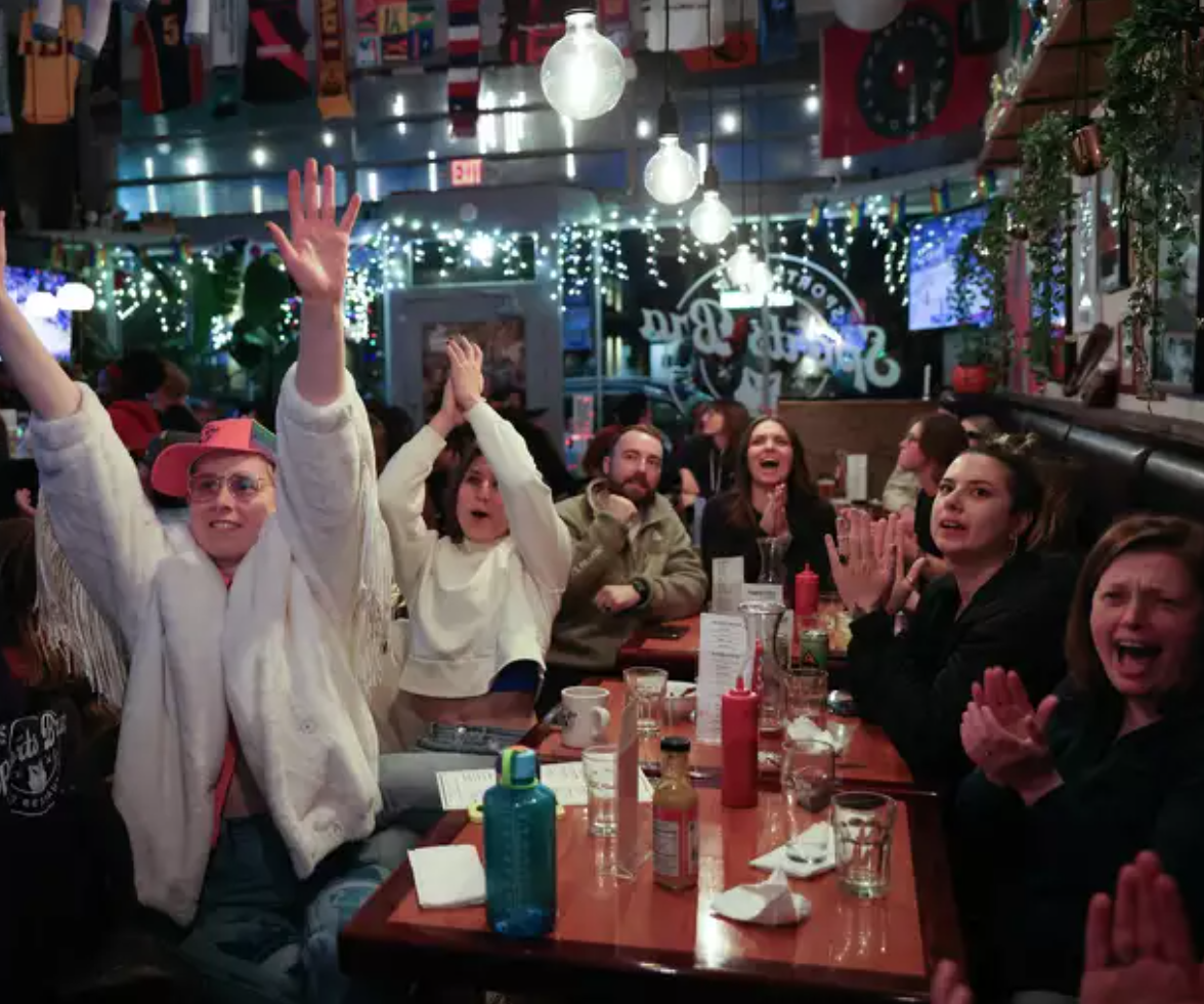 A group of people sitting at a restaurant table, cheering and clapping, with festive decorations and string lights in the background.