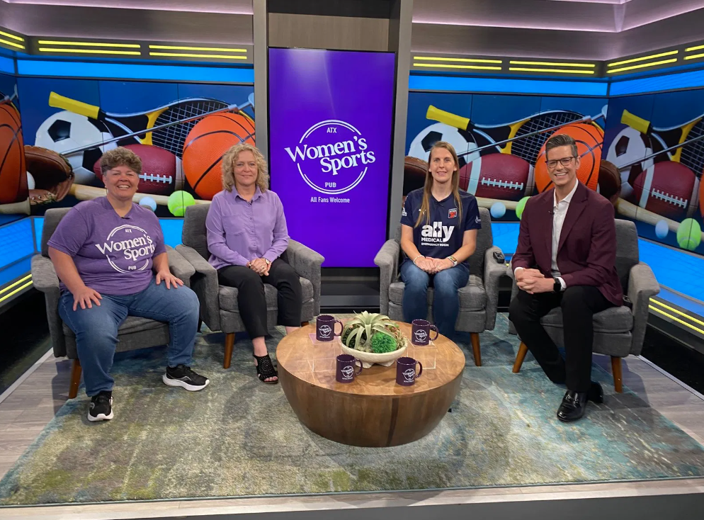 Four individuals sitting in a TV studio in front of a colorful sports-themed backdrop. Two women are wearing Women’s Sports PU shirts, and a woman is wearing a sports team jersey. A man in a suit is sitting to the right. There is a round wooden table with four mugs and a plant in the center.