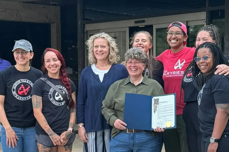 A group of women, some in athletic shirts with a rugby logo, posing together outside a building with two women in the center holding a certificate.
