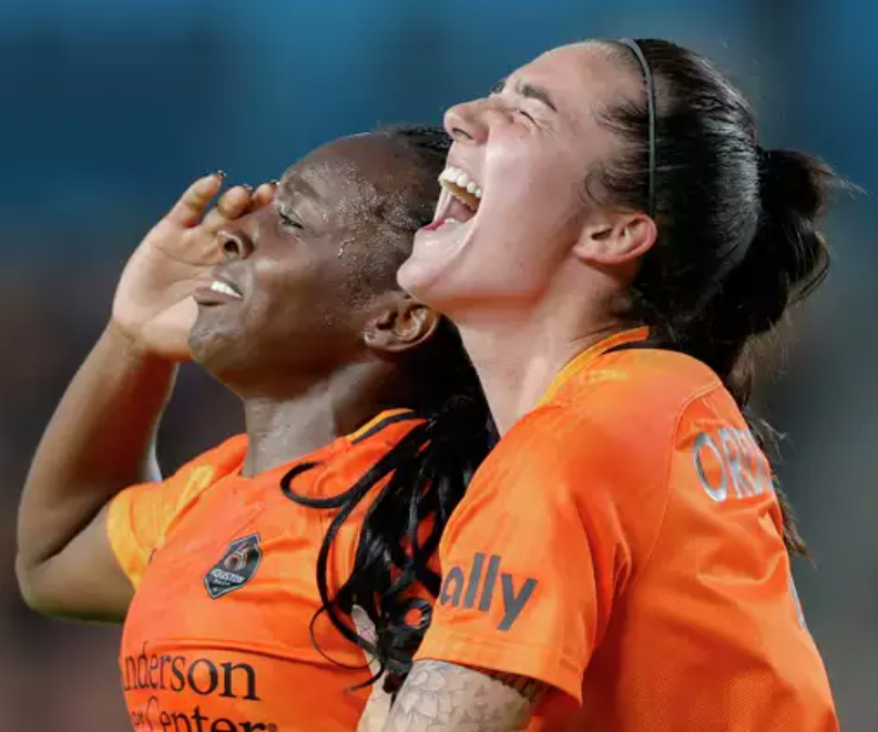 Two female soccer players in orange jerseys celebrating a victory on the field, smiling and laughing.