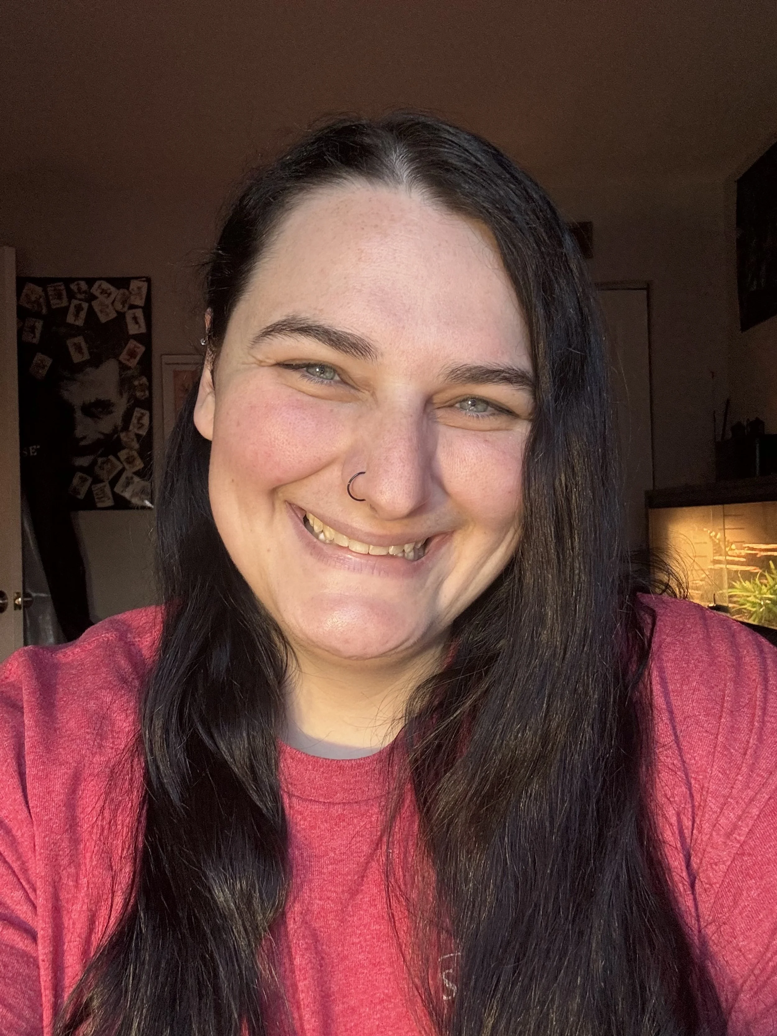A woman with long dark hair and a nose ring smiling at the camera indoors.