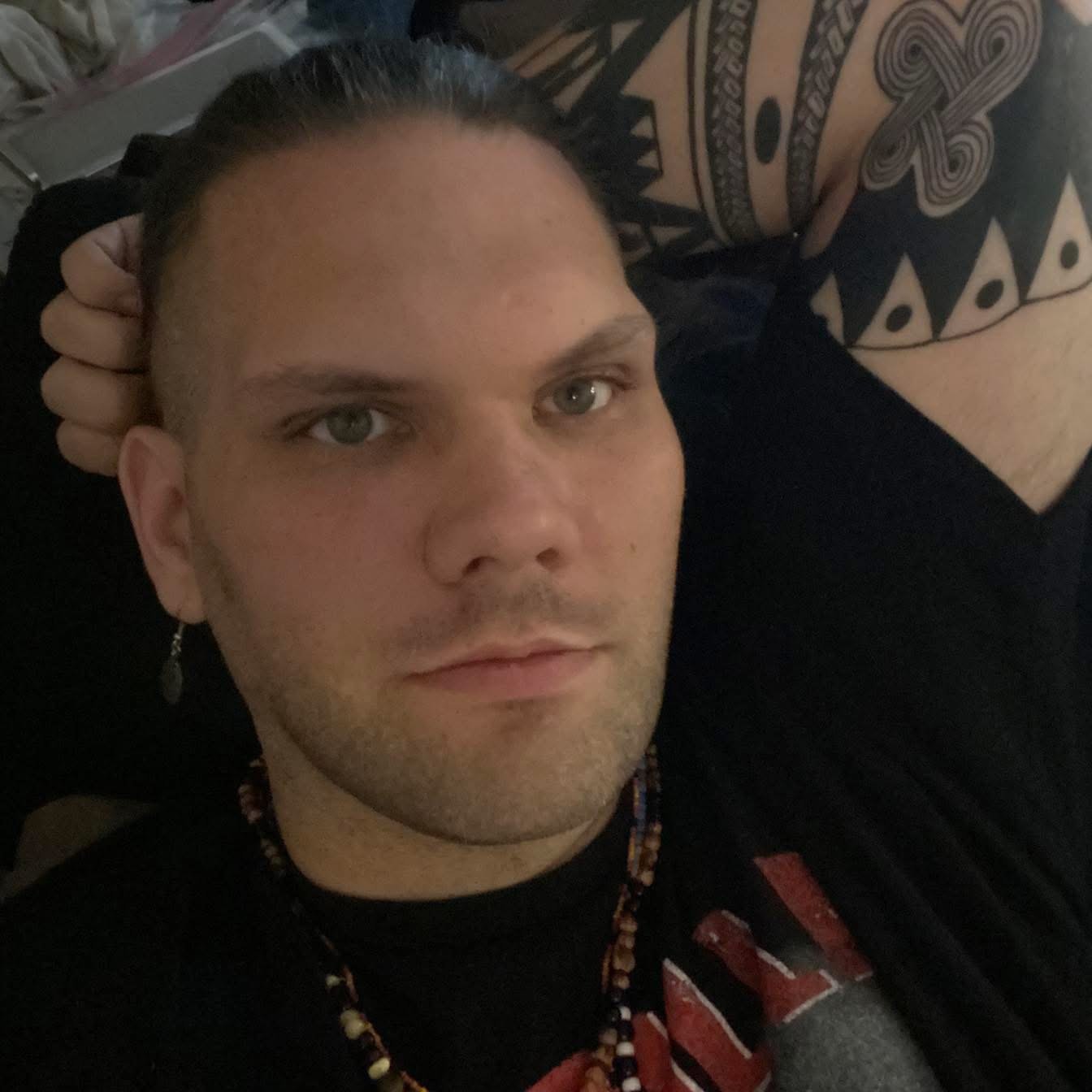 A person with light skin, short dark hair, and a beard, lying down with their head resting on a hand, and another arm showing a tribal tattoo. The person is wearing a black Eagles T-shirt, an earring, and a beaded necklace.