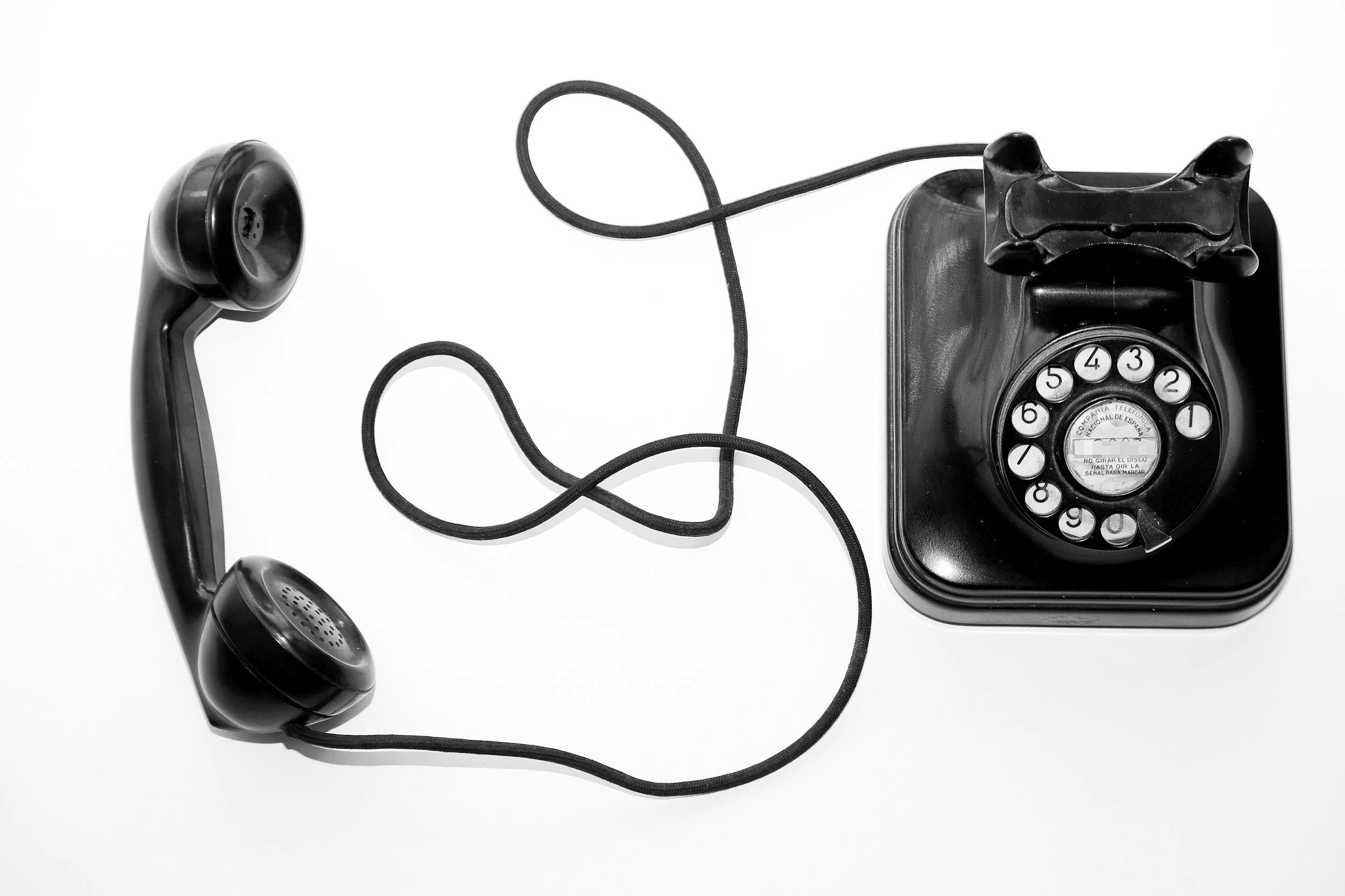 Black rotary dial telephone with cord attached, positioned on a white surface.