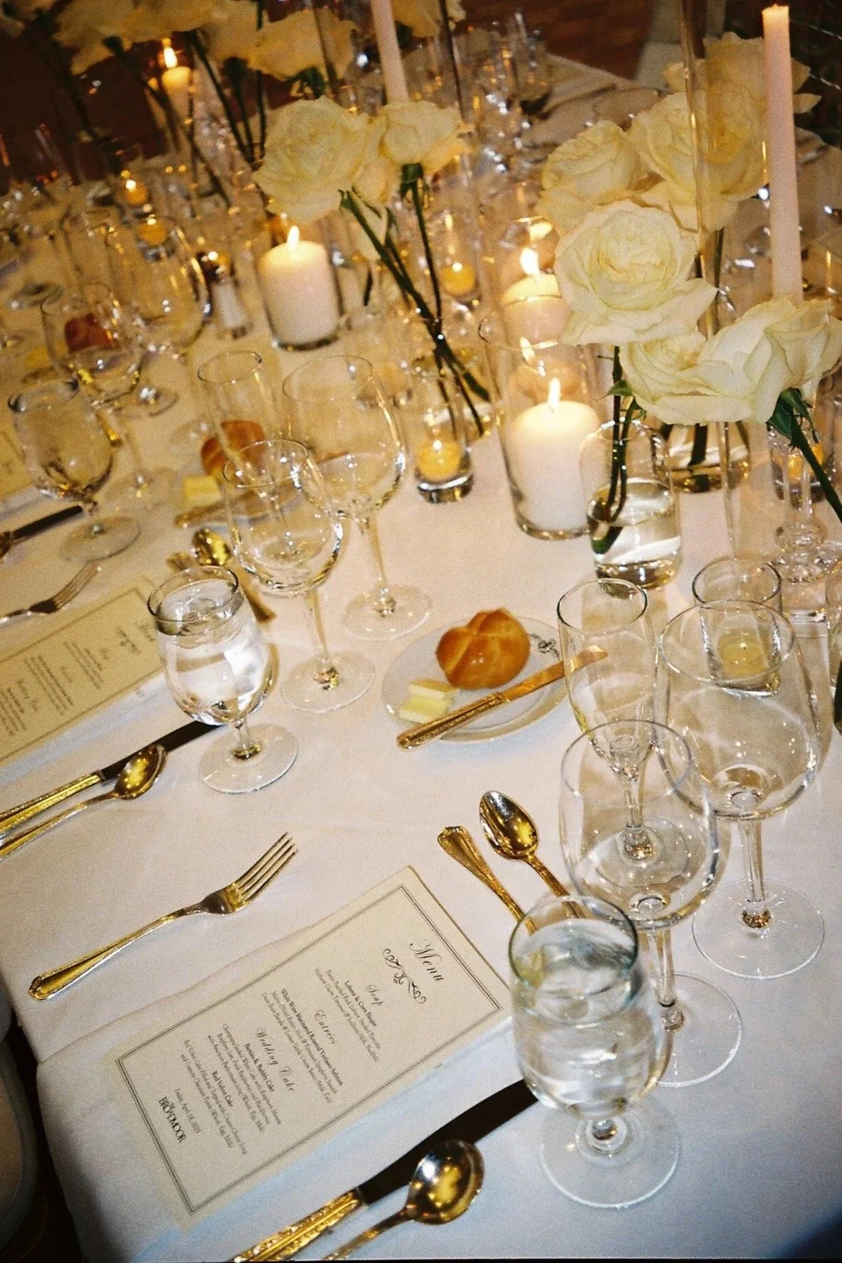 Elegant dining table setting at a formal event with white roses, candles, multiple glasses, gold utensils, and a menu on a white tablecloth.