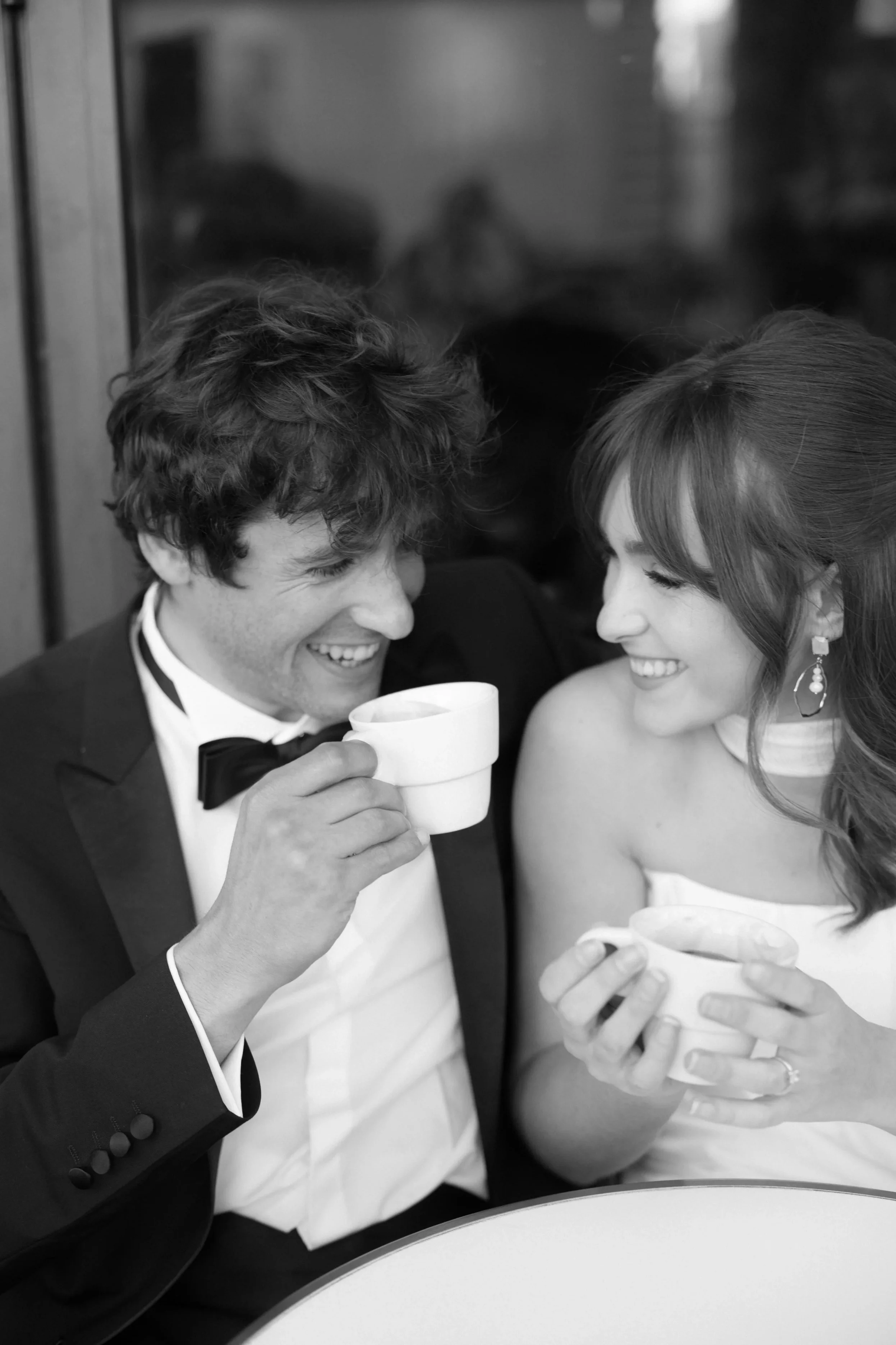 A happy couple in formal attire, smiling and sharing a moment while holding coffee cups.
