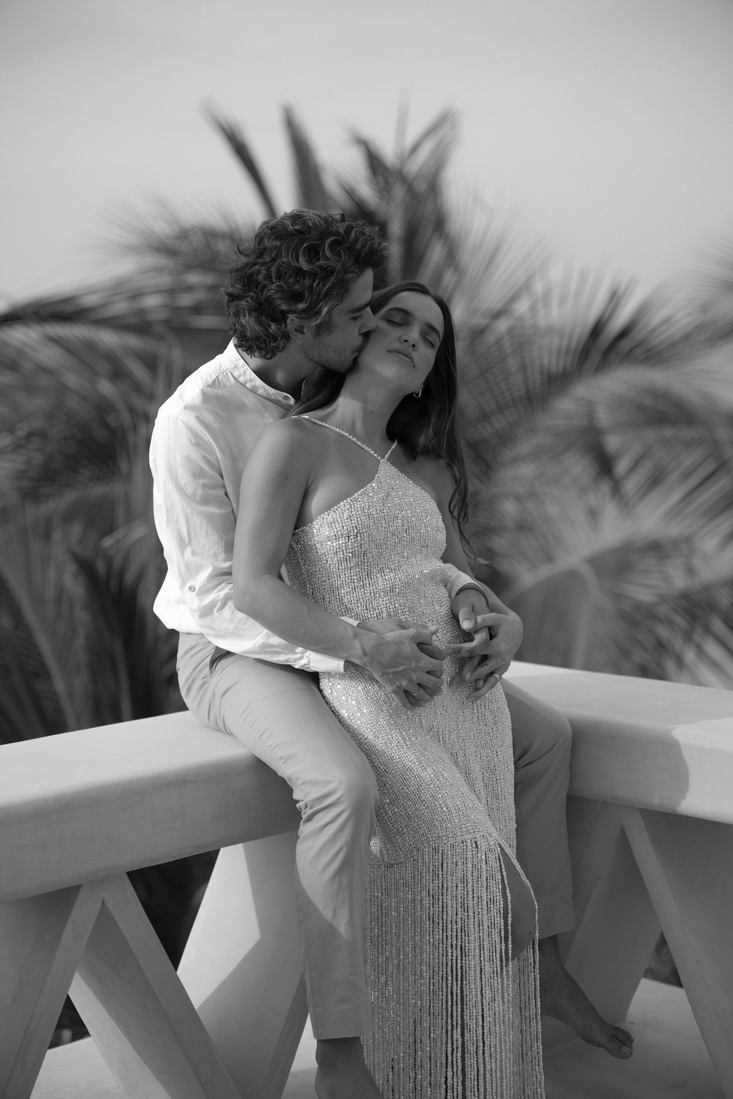 A black and white photo of a couple sitting on a bridge, with the man kissing the woman on the cheek. The woman has her eyes closed and is wearing a sparkling dress, while the man is in a light shirt and pants. There are palm trees in the background.
