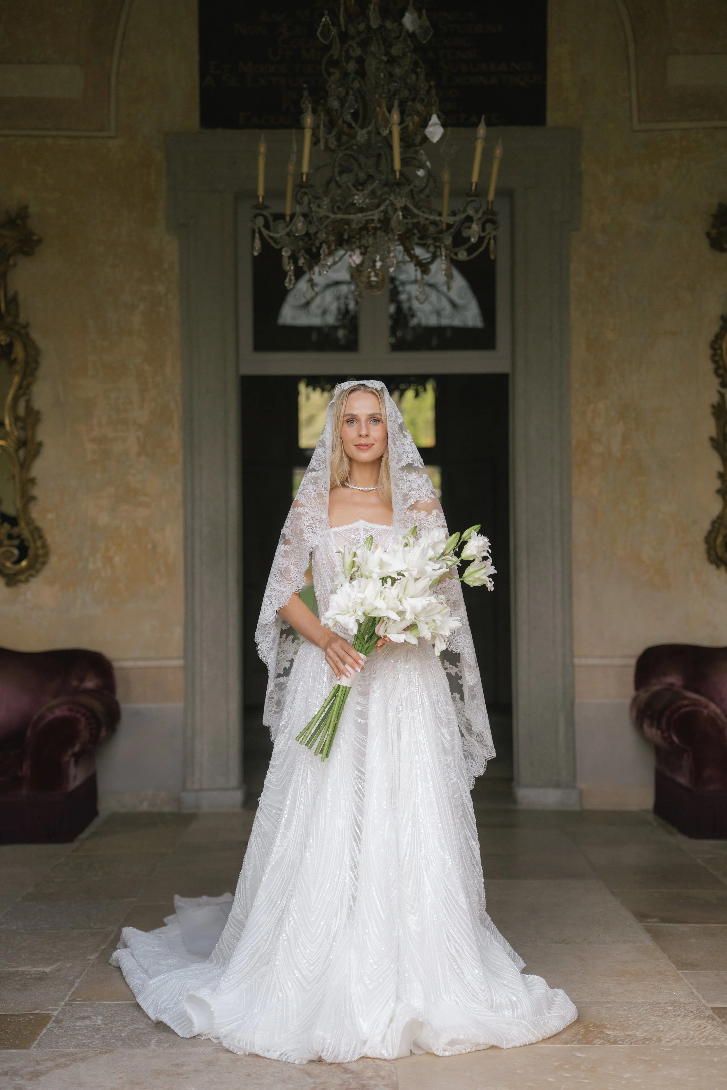 A bride in a white wedding dress holding a bouquet of white lilies, standing indoors under a chandelier.
