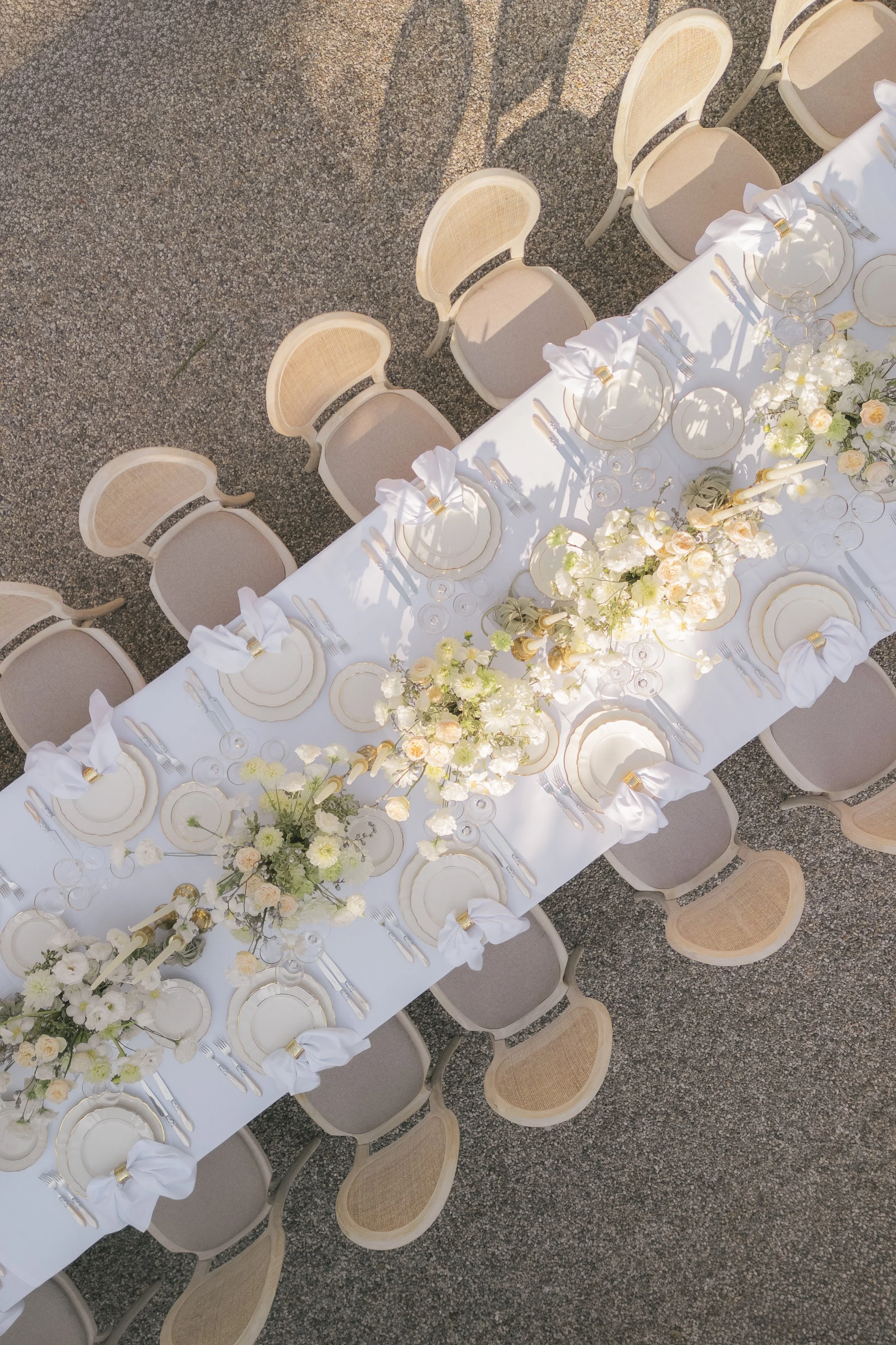 Overhead view of a long white banquet table set with dinnerware, glassware, silverware, white cloth napkins tied with gold rings, and floral centerpieces in white and pale peach colors. Beige chairs surround the table on a textured gray outdoor surfa
