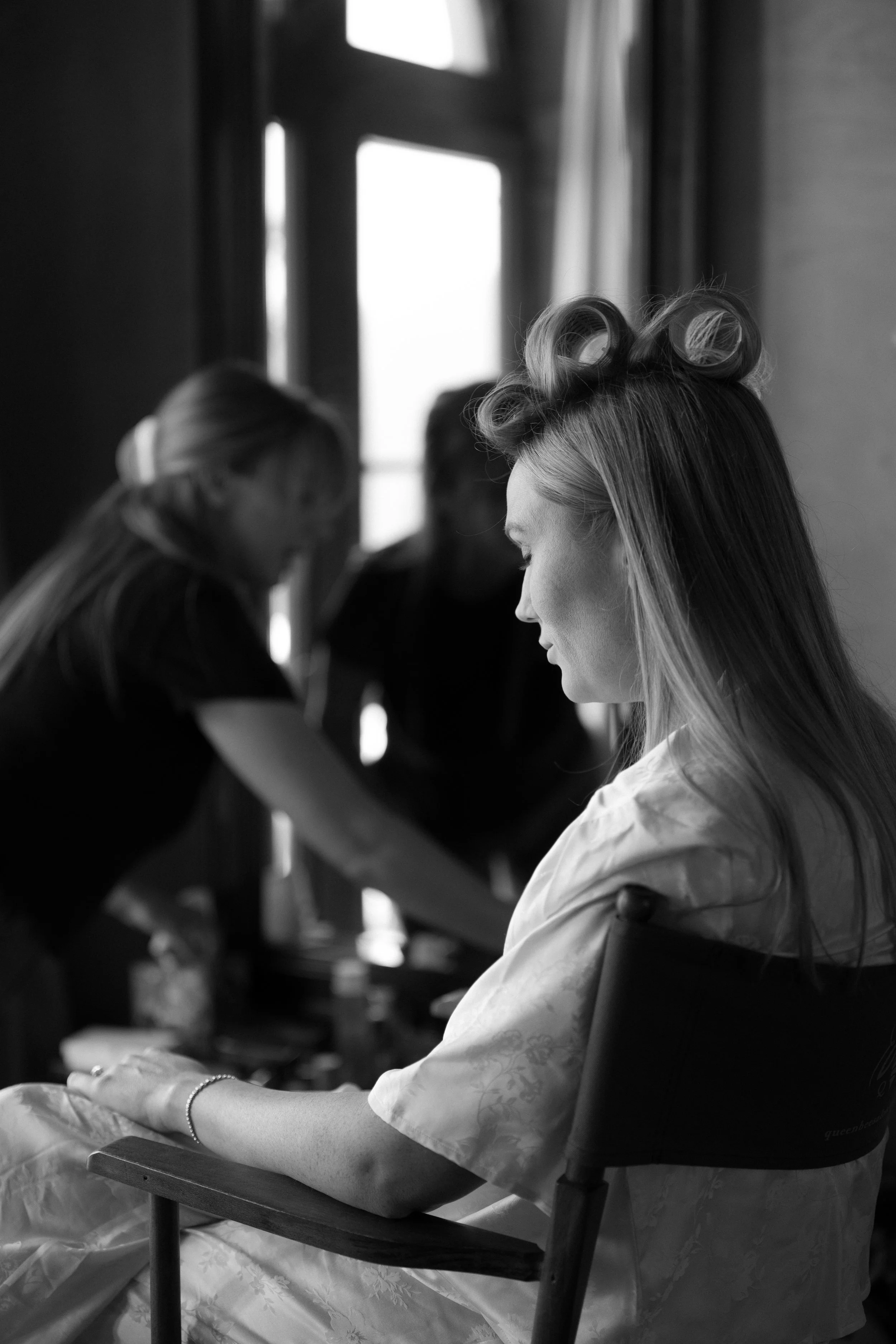 A woman with vintage hair rollers sitting in a chair, while two women in the background prepare her for an event, in front of a large window.