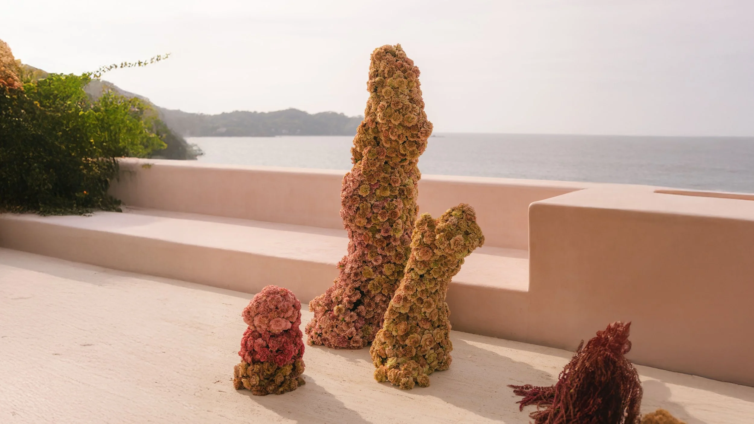 Decorative objects made of clustered dried flowers placed on a white surface outdoors, with the sea, a distant shoreline, and greenery in the background.
