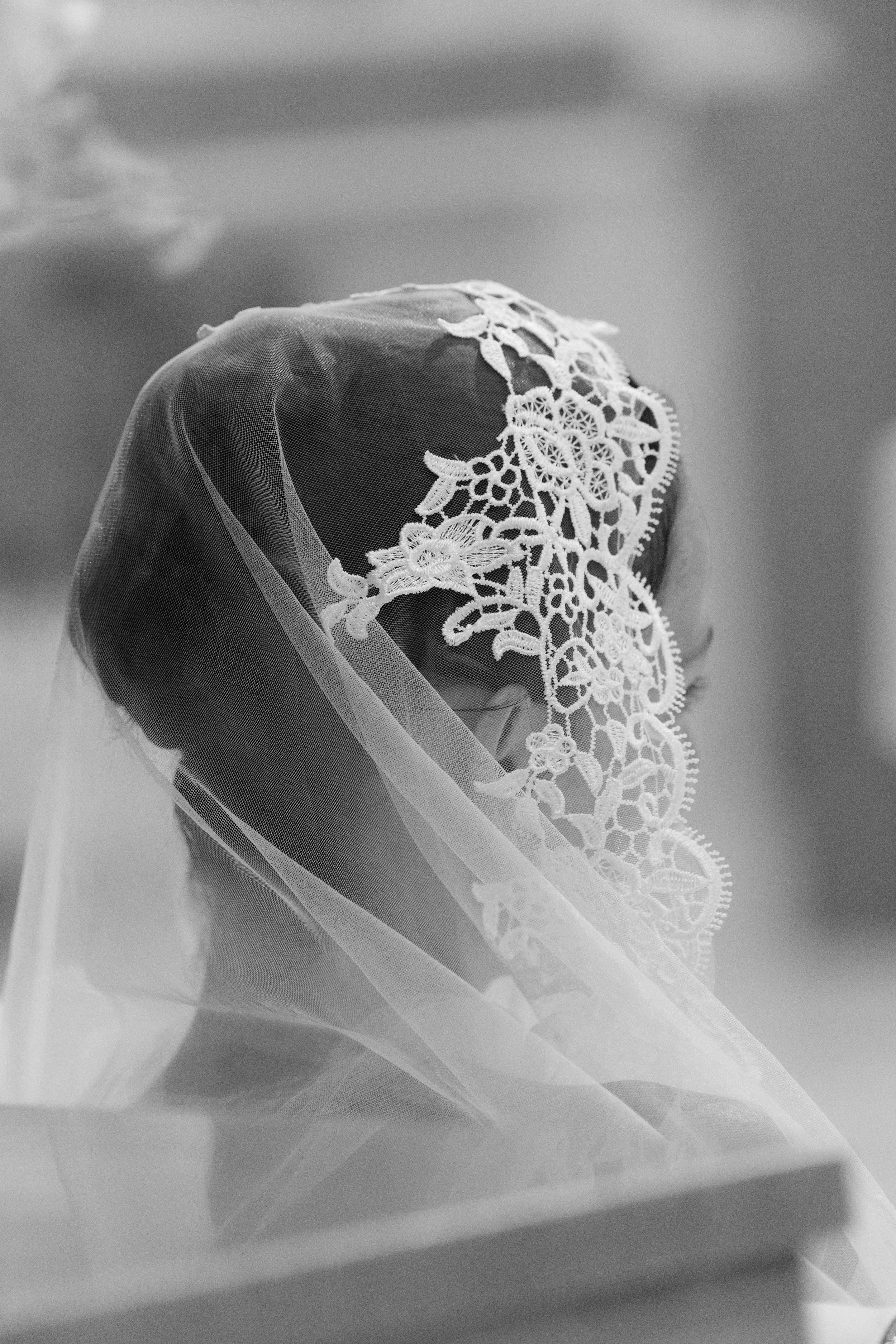 Close-up of a doll's head with a lace veil covering it, with the doll's face partially visible through the veil in black and white.