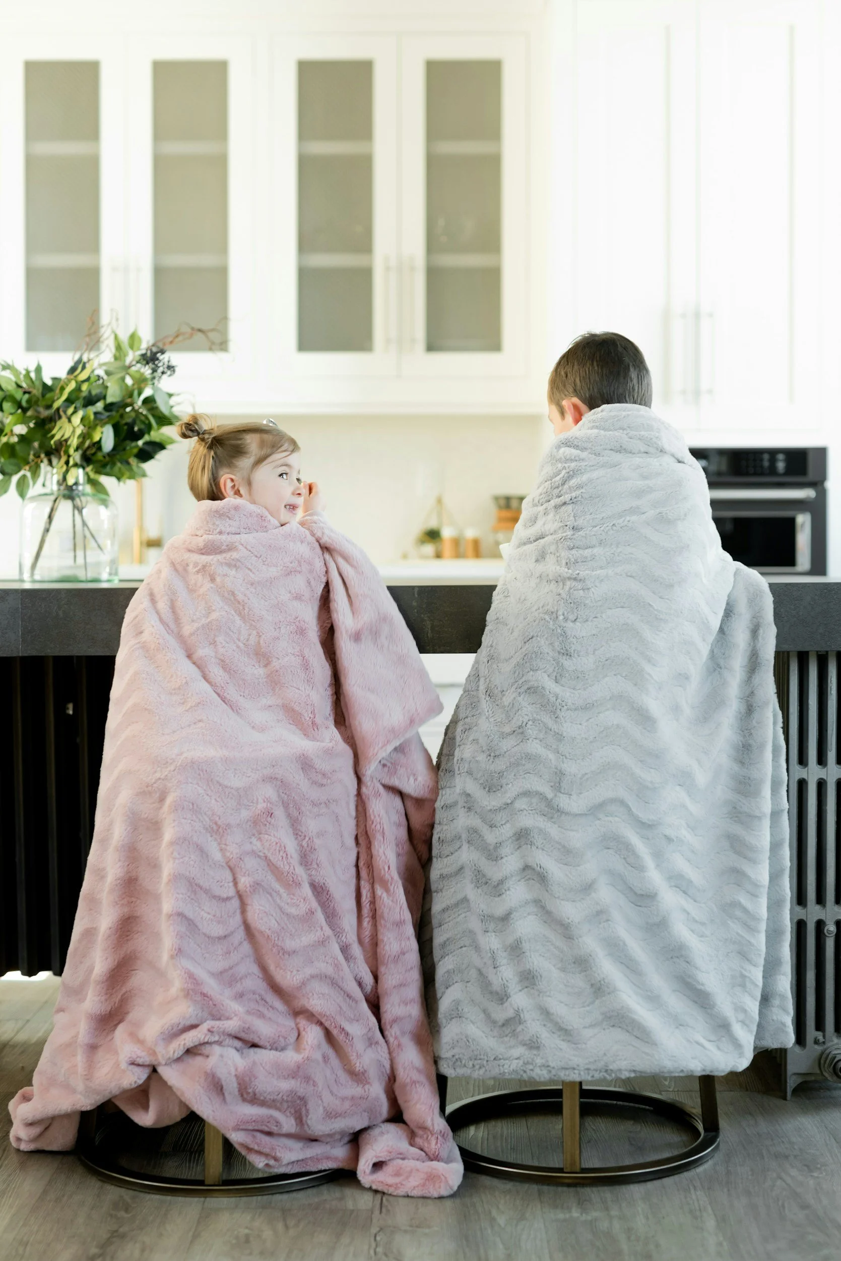 Two children, a girl and a boy, sit on chairs at a kitchen counter, wrapped in cozy blankets, engaging in conversation or sharing a moment.