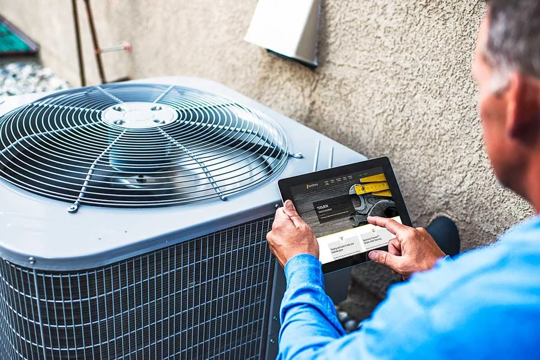 A person in a blue shirt holding a tablet with a website interface, standing next to an outdoor air conditioning unit against a beige textured wall.
