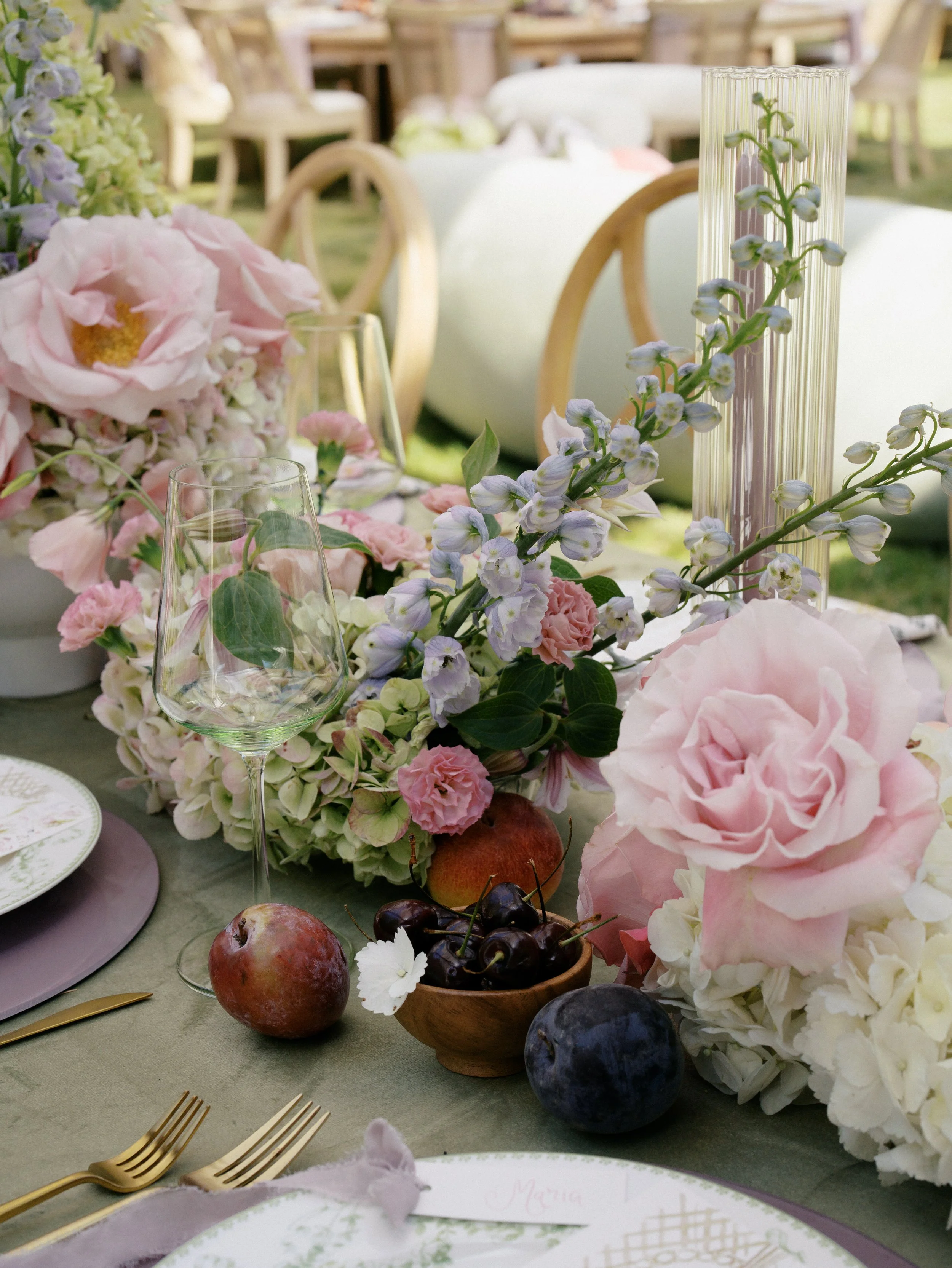 Elegant outdoor table setting with pink and white floral centerpieces, wine glasses, and gold cutlery for a celebration or wedding.